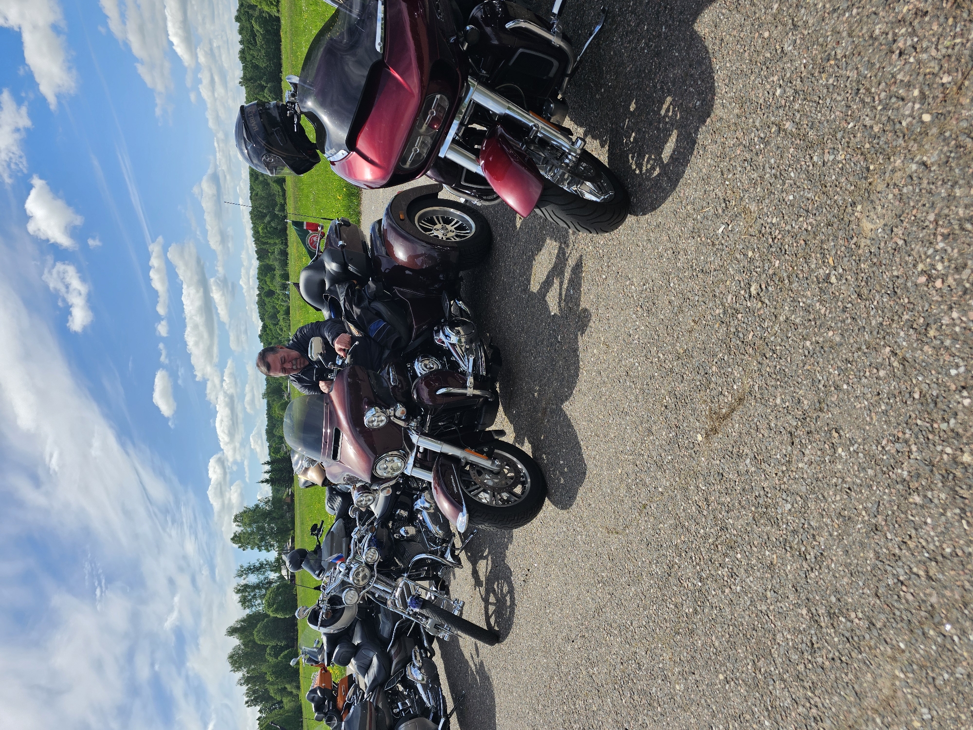 The image shows a line of motorcycles parked on a paved road. A man is sitting on one of the motorcycles, looking at the camera. The background consists of a green field and a blue sky with clouds. The location is possibly near Borodinskogo Muzeya, Russia, given the presence of motorcycles suggesting a gathering or event.