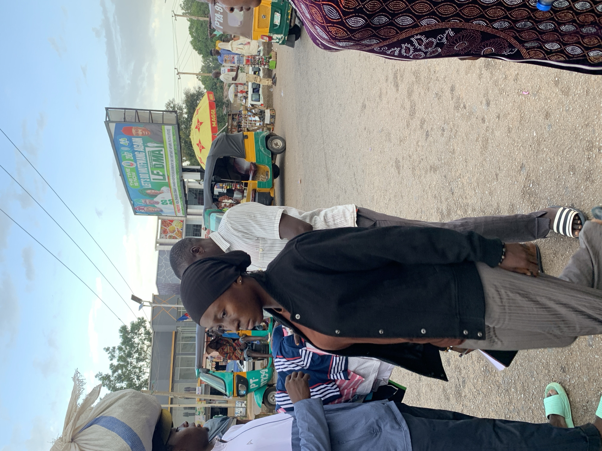 The image depicts a street scene in Jos, Nigeria, likely near a busy intersection or market. The sky is partly cloudy, suggesting it is either morning or late afternoon. In the foreground, a young woman wearing a black headscarf and jacket is looking to her left, with a man in a white and blue striped shirt standing behind her. To her right, another person in a patterned purple garment is partially visible.
In the middle ground, a yellow and green auto-rickshaw, commonly known as a 'Keke Napep', is parked, its driver out of frame. Another auto-rickshaw is visible further down the street. There are several stalls and shops lining the roadside, some with umbrellas providing shade. A large billboard with political advertisements is prominently displayed in the background. One advertisement features images of politicians and the text "THE TIME IS NOW. LET'S DO MUTTWANG AGAIN. LE DIMA". Another part of the billboard shows the 'MAGGI' logo on a yellow umbrella.
The ground is paved with asphalt and gravel, typical of a road in a developing urban area. People are casually dressed, with one person in the foreground wearing light blue slide sandals. The overall impression is one of everyday life and commerce in a bustling Nigerian city.