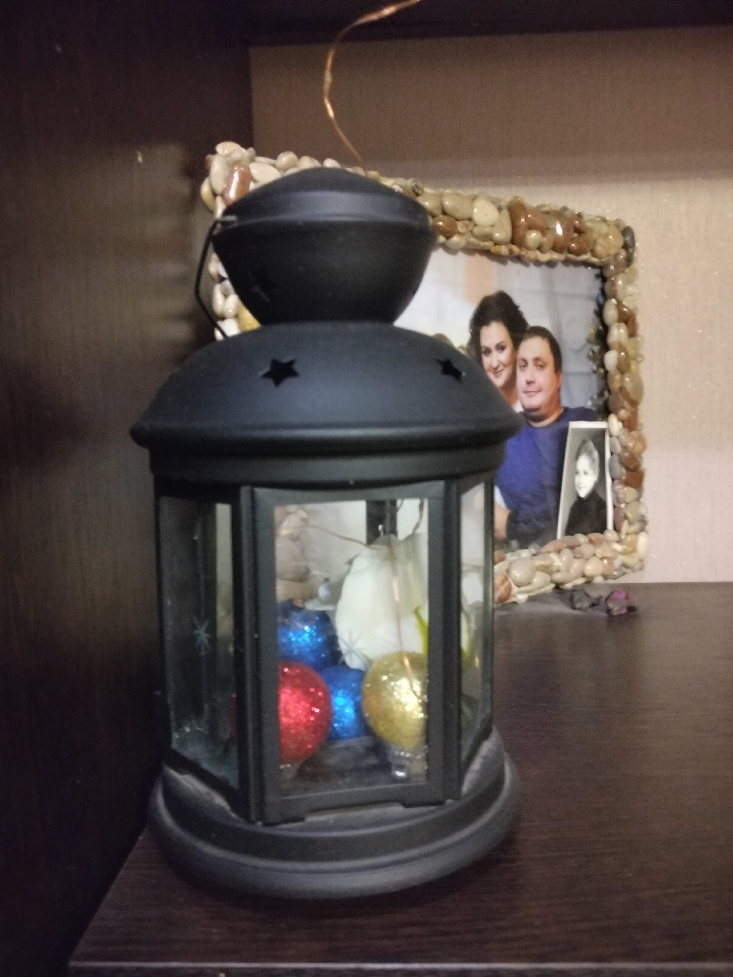A black lantern with glass panels sits on a dark wooden shelf. Inside the lantern, there are decorative ornaments, including a red and a gold glittery ball, and some white objects. Behind the lantern, a framed photo is visible. The frame is decorated with small stones. The photo itself shows a couple, with the woman on the left and the man on the right. A smaller inset picture of a child is also within the frame. The background appears to be a wall. The overall setting suggests a domestic environment, possibly a living room or shelf in a home. There is no indication of the location being in Nizhnedonskoy, Russia.