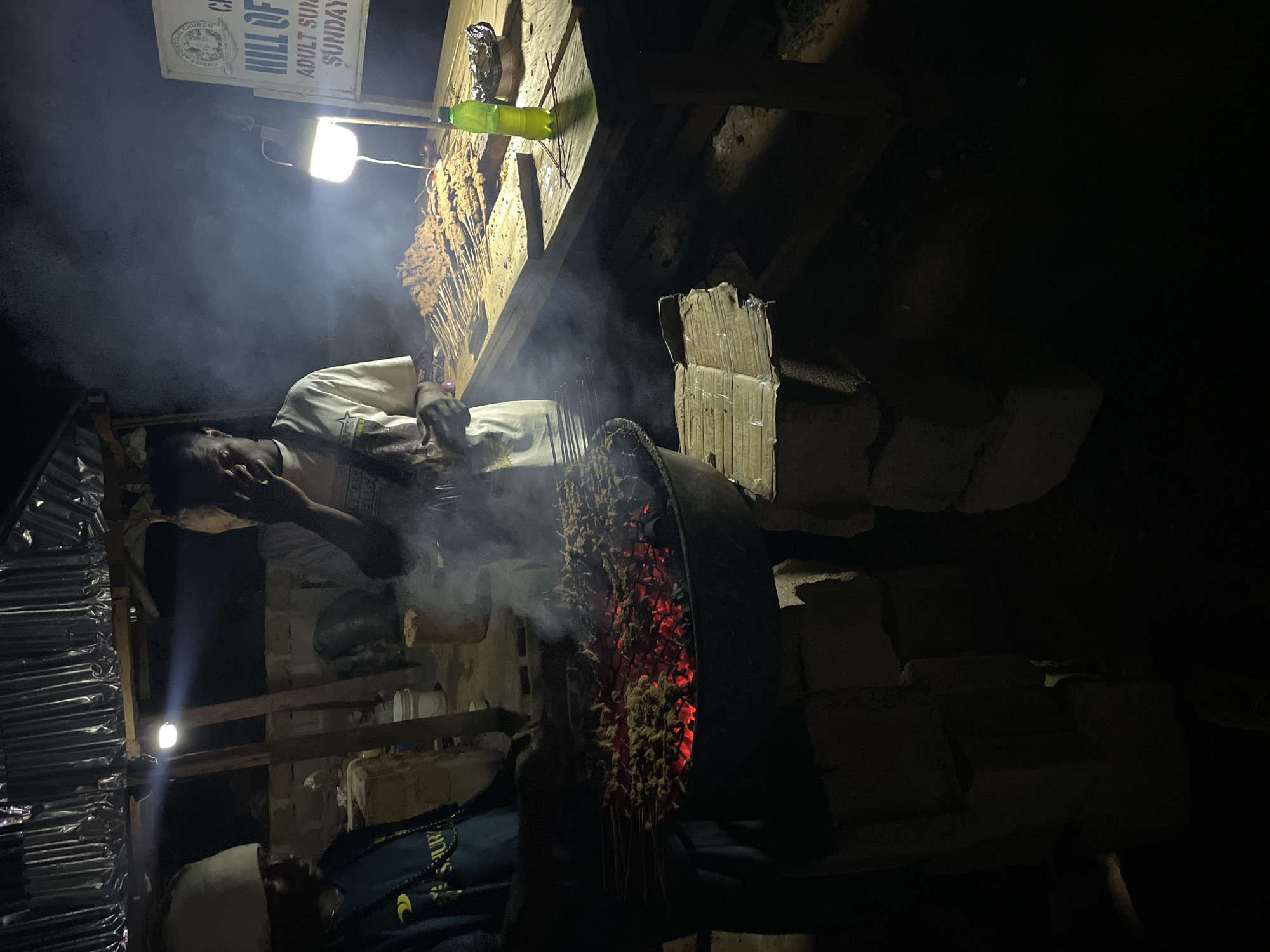 In Durimi, Nigeria, a man tends to a barbeque grill loaded with skewers, visible through a haze of smoke and glowing embers. The grill stands on a makeshift structure of cinder blocks. The man wears a light-colored shirt. A wooden table to the left holds more skewers. A light illuminates the table. A sign reading "HILL OF ADULT SUNDAY" is visible above the table. The surrounding environment is dark.
