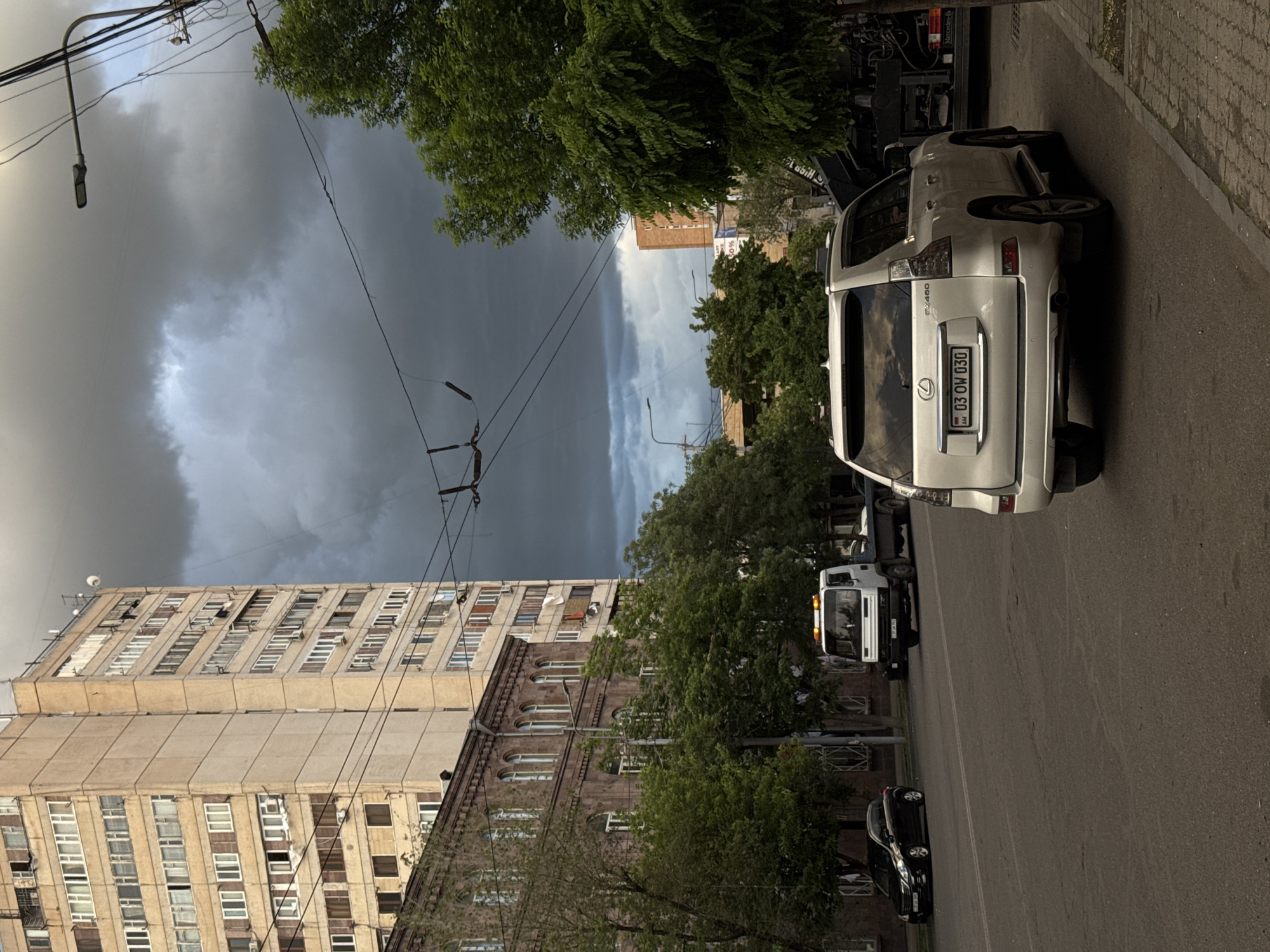The image depicts a street scene in Yerevan, Armenia, likely on or near Aram Street, based on the architectural style of the buildings. The sky is dominated by dark, ominous storm clouds. A light-colored Lexus SUV, license plate "03 OW 030", is parked on the right side of the street, facing away from the camera. Several other vehicles are parked along the street further away, including a white truck. Tall trees line the street, partially obscuring the buildings. Power lines crisscross overhead. The buildings on the left include a tall, Soviet-era apartment building and a brick building with arched windows. The street is paved and appears to be in urban neighborhood. The scene suggests an approaching storm.