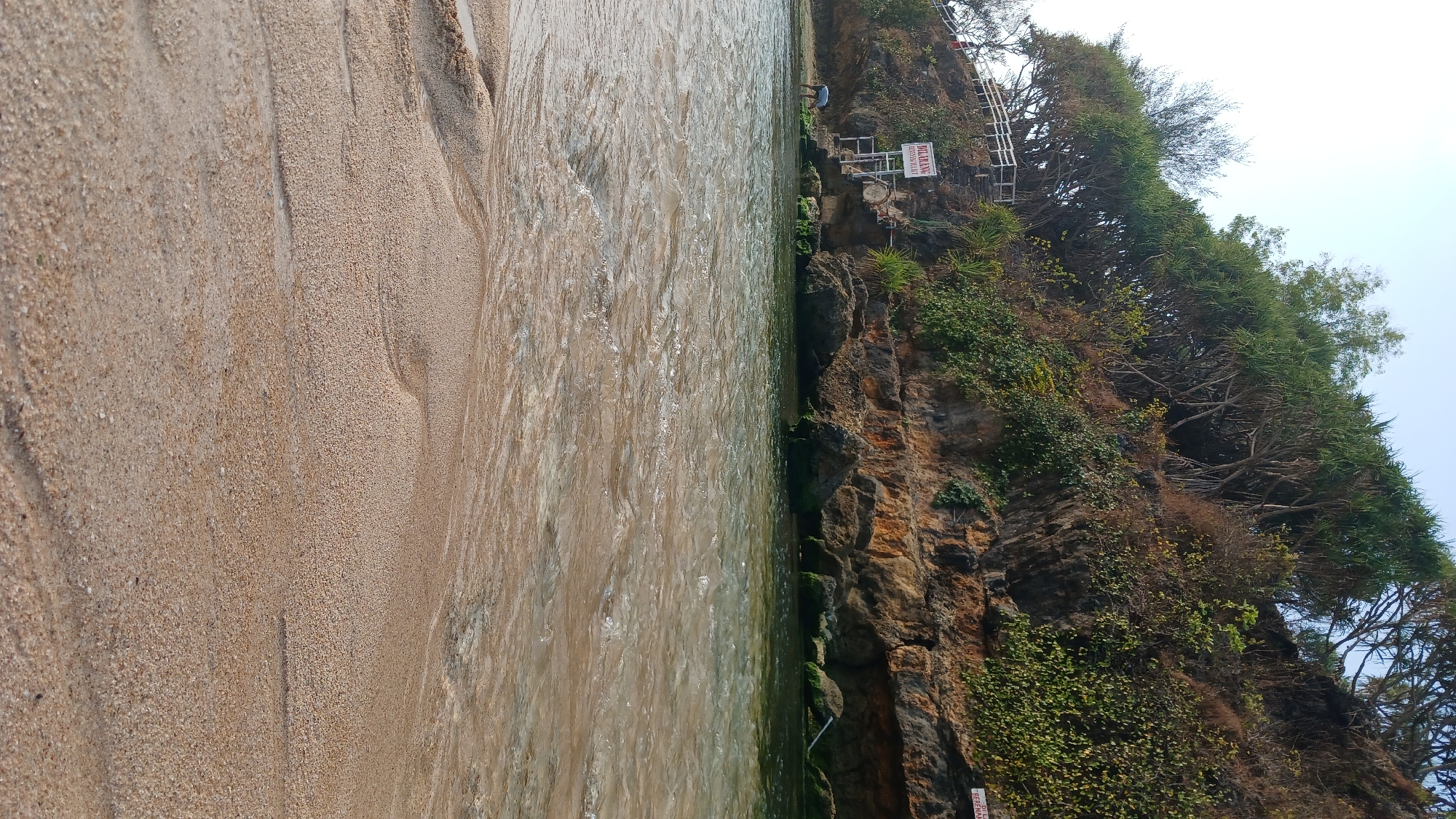 The image captures a serene beach scene in Tanjungsari, Indonesia. The foreground is dominated by wet, light-brown sand with gentle ripples left by the receding tide. A person, appearing to be male, is visible in the mid-ground, bent over near the water's edge. In the background, a rocky cliff face is covered with lush green vegetation and trees, creating a natural backdrop. Signs are posted on the cliffside, one of which reads "DILARANG BERENANG" in red lettering, indicating a prohibition against swimming. The water is clear and shallow, with sunlight reflecting off its surface, creating a shimmering effect. The sky above is a pale blue with a slight haze.