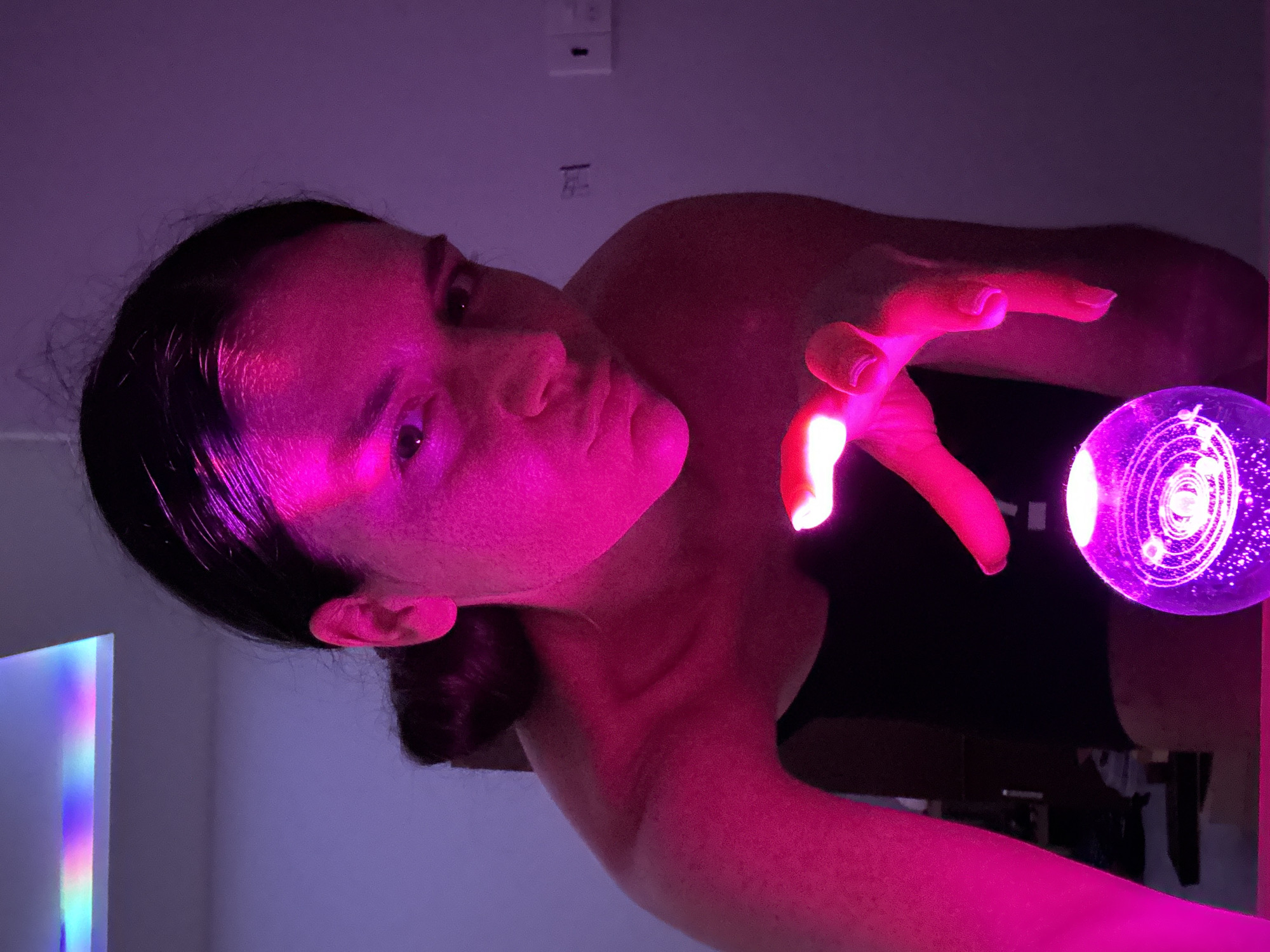 A young woman with dark hair pulled back in a bun is leaning forward, her face illuminated by a vibrant pink light. She is looking towards the viewer with a neutral expression. Her right hand is extended over a clear, spherical object that appears to be a snow globe or decorative orb containing a model of the solar system. The orb is also glowing with a pinkish-purple light, highlighting the planetary models within. The background is a softly lit room with a purple hue. A strip of rainbow-colored light is visible on the ceiling in the upper left corner of the image, suggesting an artificial light source with color-changing capabilities. The woman is wearing a dark top. The scene suggests a playful or mystical moment captured in an indoor setting in Mũi Né, Vietnam.