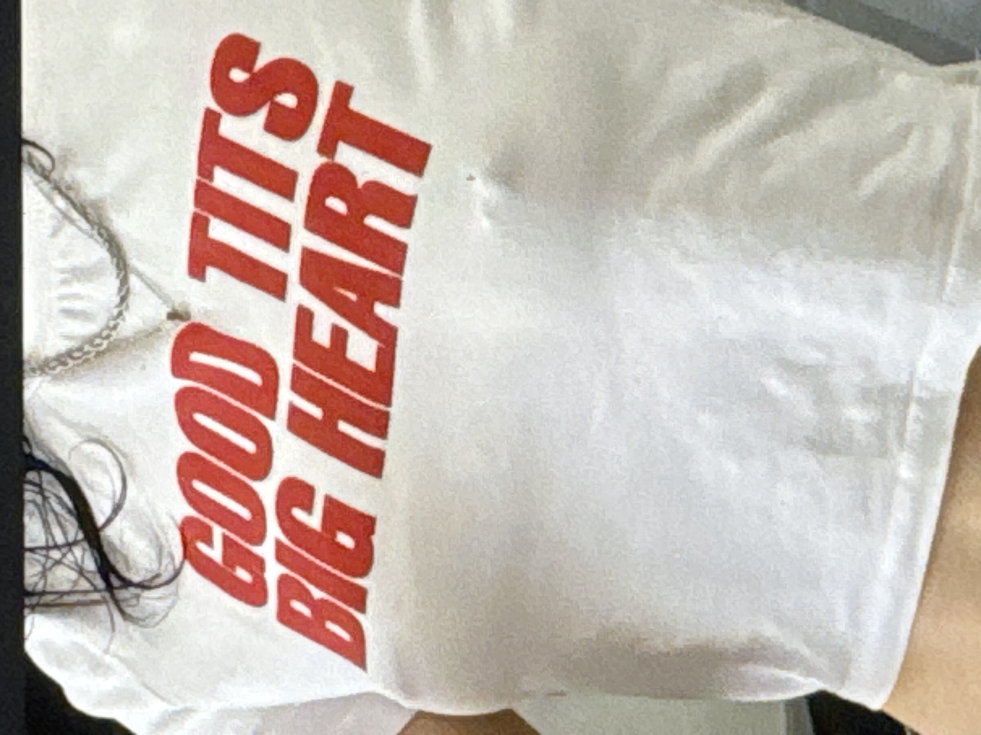 A close-up shot shows a person wearing a white t-shirt with red lettering. The text on the shirt reads "GOOD TITS BIG HEART". The person is wearing a silver chain necklace over the t-shirt. The background is blurred and out of focus. The image does not contain any discernible location details for Kuta Selatan, Indonesia.
