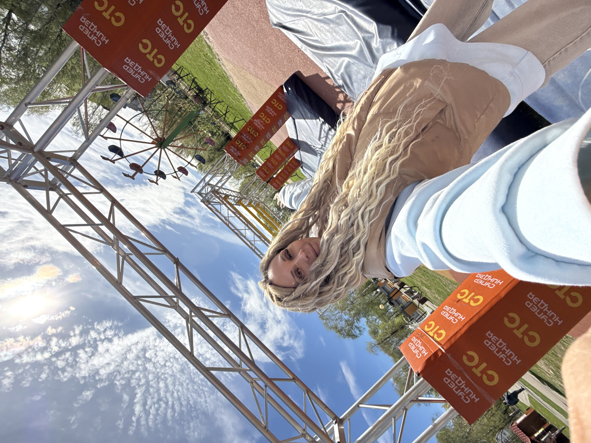 The image shows a woman at an outdoor event in Unknown, Unknown. The camera's point of view is low, tilted upwards towards the sky, giving an unusual perspective. The woman is in the foreground, wearing a light blue hoodie and a tan jacket, with long blonde dreadlocks.
In the background, an amusement park ride, possibly a swing ride, can be seen. Metal scaffolding and orange signs with the text "CTC" and the words "Супер Ниндзя" in Russian are also prominent in the frame. The sky is bright blue with some wispy clouds, and the sun's glare is visible in the upper left corner. There is also some greenery, possibly grass and trees, visible in the background. The woman seems to be posing for the camera, with a slight smile on her face.