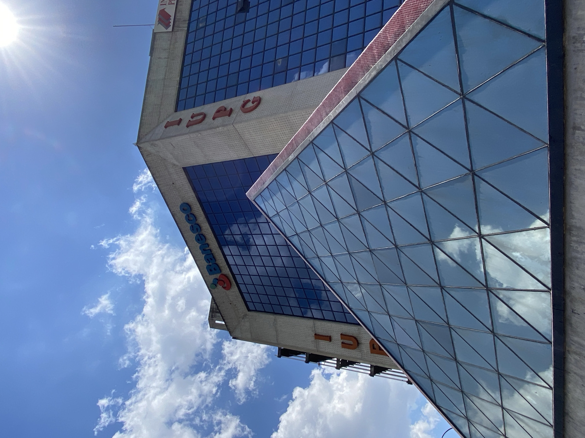 An upward-angled view depicts a modern urban building complex under a bright, partly cloudy sky in Chacao, Caracas, Venezuela. The foreground is dominated by a geometrically faceted glass pyramid structure, likely an atrium or skylight, composed of dark-framed triangular panes reflecting the blue sky and white clouds. A reddish-brown textured strip runs along the upper right edge of this pyramid. Behind and to the right, a multi-story building features a concrete frame and extensive dark blue-tinted glass windows, also reflecting the sky. Prominent signage includes the "Banesco" logo in blue text with a red and orange emblem on the upper facade, and the red capital letters "IUPG" oriented vertically along the building's corner. A partial sign showing "IN" and "PR" in red is visible on the far upper right. An external metal stairwell or fire escape is present on the left side of the concrete building. A strong sunburst is visible in the upper right quadrant of the sky, indicating direct sunlight. No people, specific actions, or interactions are present in the static scene. The building is identifiable as the Banesco building associated with the Instituto Universitario Politécnico Gran Colombia (IUPG) on Avenida Francisco de Miranda.
