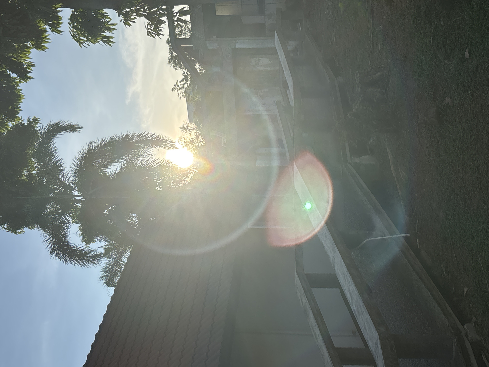 The image, likely taken in the city of บ้านบางรักษ์, Thailand, is an upward angle shot with the sun directly in the frame, creating significant lens flare. The foreground shows a red-tiled roof and a large palm tree to the left. In the background, there's a structure that appears to be a concrete building or raised walkway. A portion of a grassy area borders the walkway. The sky is partially visible behind the structures and trees. The strong sunlight and lens flare obscure many details.