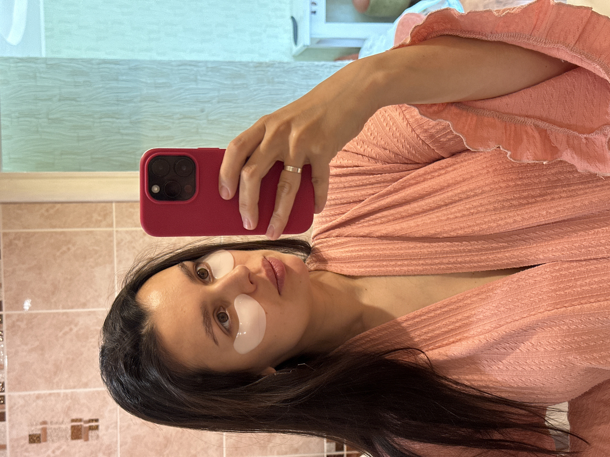 A woman is taking a selfie in a bathroom. She is wearing eye masks and a peach-colored robe. She is holding a red smartphone in her right hand. She is wearing a ring on her ring finger. The bathroom has peach-colored tiles. The location is potentially Chaikovsky, Russia, but there is no definitive proof.