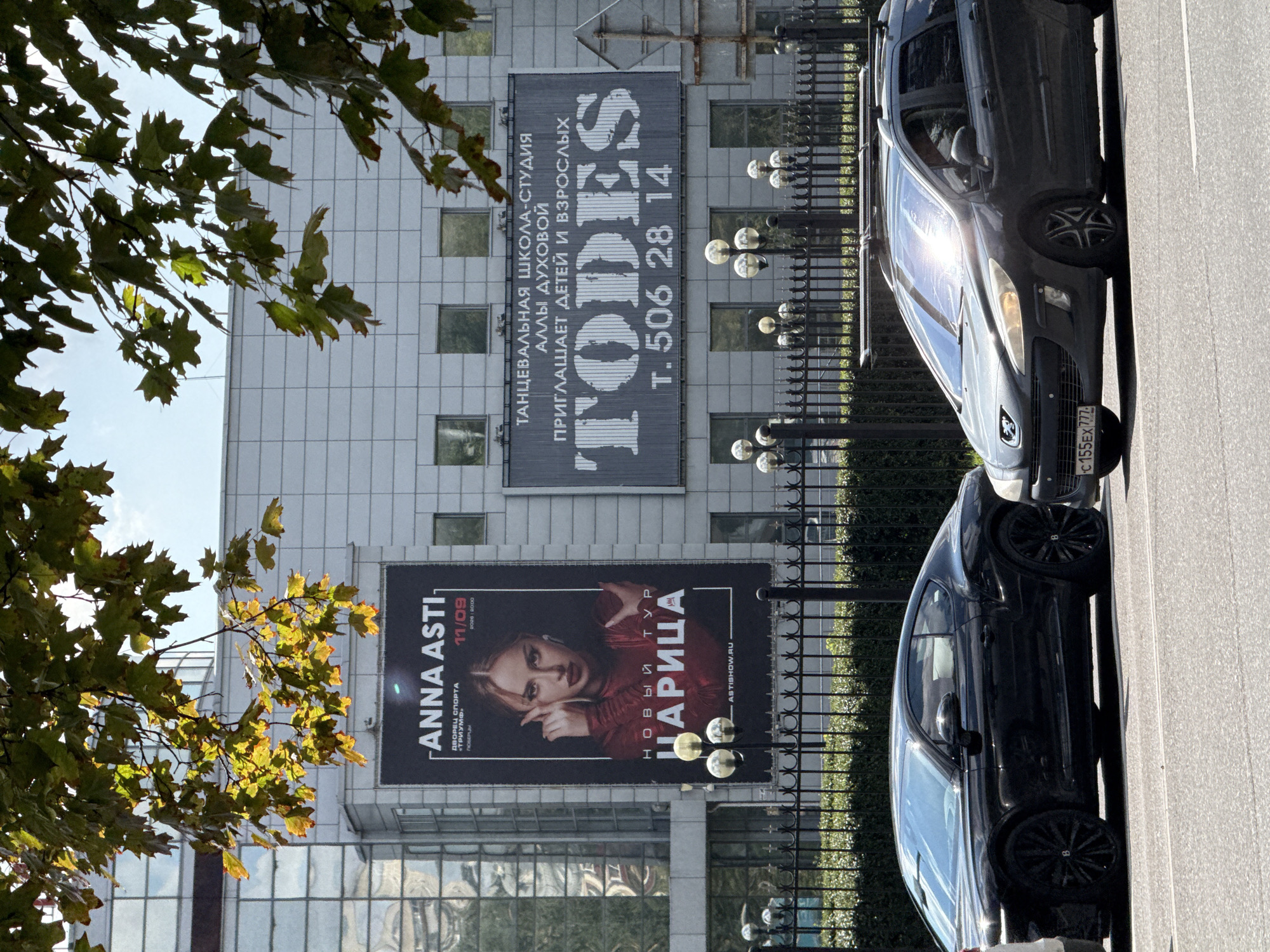 In the city of Tomilino, Russia, on a bright, sunny day, two cars are parked side-by-side on a street in front of a large, modern building. The building features a white tiled facade with numerous windows and two prominent billboards. The billboard on the left displays a poster for a concert by "ANNA ASTI" with the title "НОВЫЙ ТУР ЦАРИЦА" (New Tour Empress) and the date 11/09. The billboard on the right advertises a dance studio named "TODES," including a phone number. In the foreground, a black coupe-style car, possibly a Bentley, is parked on the left, and a dark-colored station wagon is parked on the right. A black metal fence runs along the front of the property, behind which several ornate lamp posts are visible. Lush green foliage from tree branches hangs overhead, partially obscuring the top of the building and casting dappled shadows.