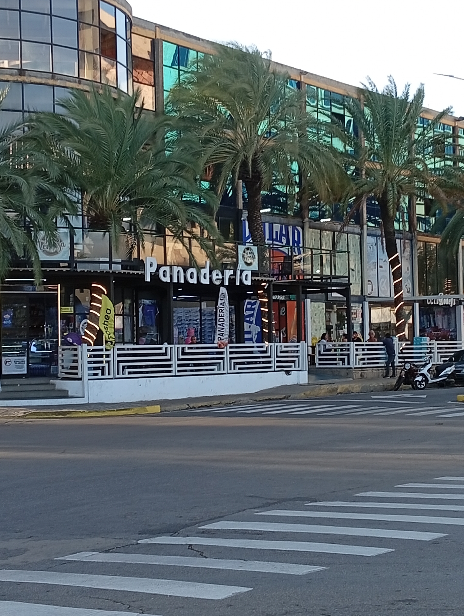 The image shows a street scene in Juan Griego, Venezuela, with a "Panaderia" (bakery) visible in the background. The bakery has outdoor seating enclosed by a white fence. People are seated at tables. A person stands near parked scooters. A crosswalk is in the foreground of the scene, with an asphalt road. The building behind the bakery has blue-tinted glass windows, and palm trees are planted around the building. "LEAD" is visible on a sign. The sky is bright and clear.