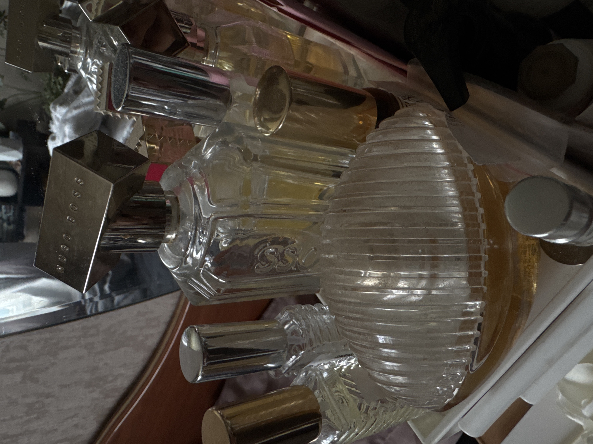 The image presents a close-up, tilted shot of a cluttered collection of perfume bottles, mainly glass, in a variety of shapes and sizes. Several bottles feature metallic accents, such as silver or gold caps and spray mechanisms. Brands visible include "HUGO BOSS" and "SSC." The background is generally blurred, indicating a shallow depth of field, but glimpses of furniture, possibly a tabletop and a drawer, suggest an indoor environment. The scene appears to be a private residence, given the personal nature of the objects. No information is present that indicates a connection with Izhevsk, Russia.
