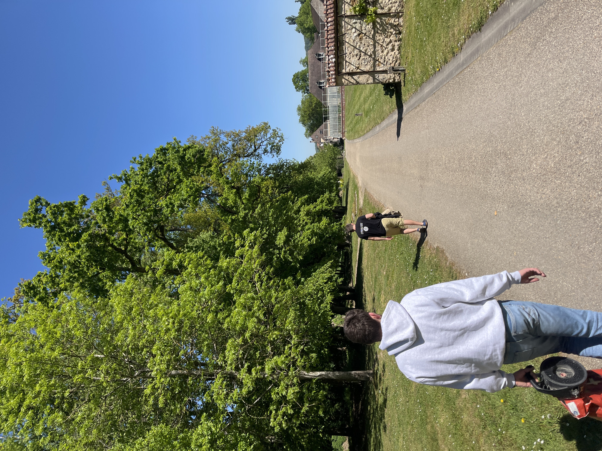 The image captures a sunny outdoor scene in Saint-Rémy-lès-Chevreuse, France, featuring two individuals on a pathway. One person, seen from the back, is wearing a light gray hoodie and jeans, and is holding a red and black lawn care device. The other individual, further down the path, wears a black t-shirt and khaki shorts. The pathway stretches towards a building with a greenhouse. Lush green trees border the path, and the sky is clear and blue.