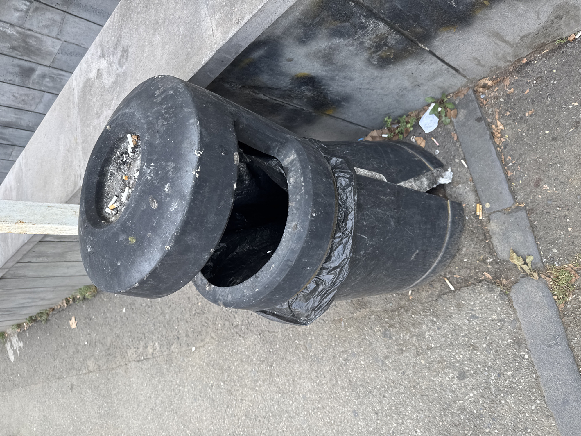 An outdoor ashtray bin, with a black plastic body, is located on a paved sidewalk next to a concrete wall. The bin has two openings, one on top and one in the middle. The top opening serves as an ashtray and contains several cigarette butts and ash. The middle opening is a larger receptacle, and a black plastic bag is visible inside, partially pulled out. The bin is damaged, with a crack running down its side. The sidewalk is made of asphalt and appears dirty, with scattered cigarette butts and dried leaves. The concrete wall behind the bin is textured and shows signs of wear and some discoloration. The overall scene suggests an urban environment, possibly in a public area where smoking is permitted. The lighting indicates it is daytime.