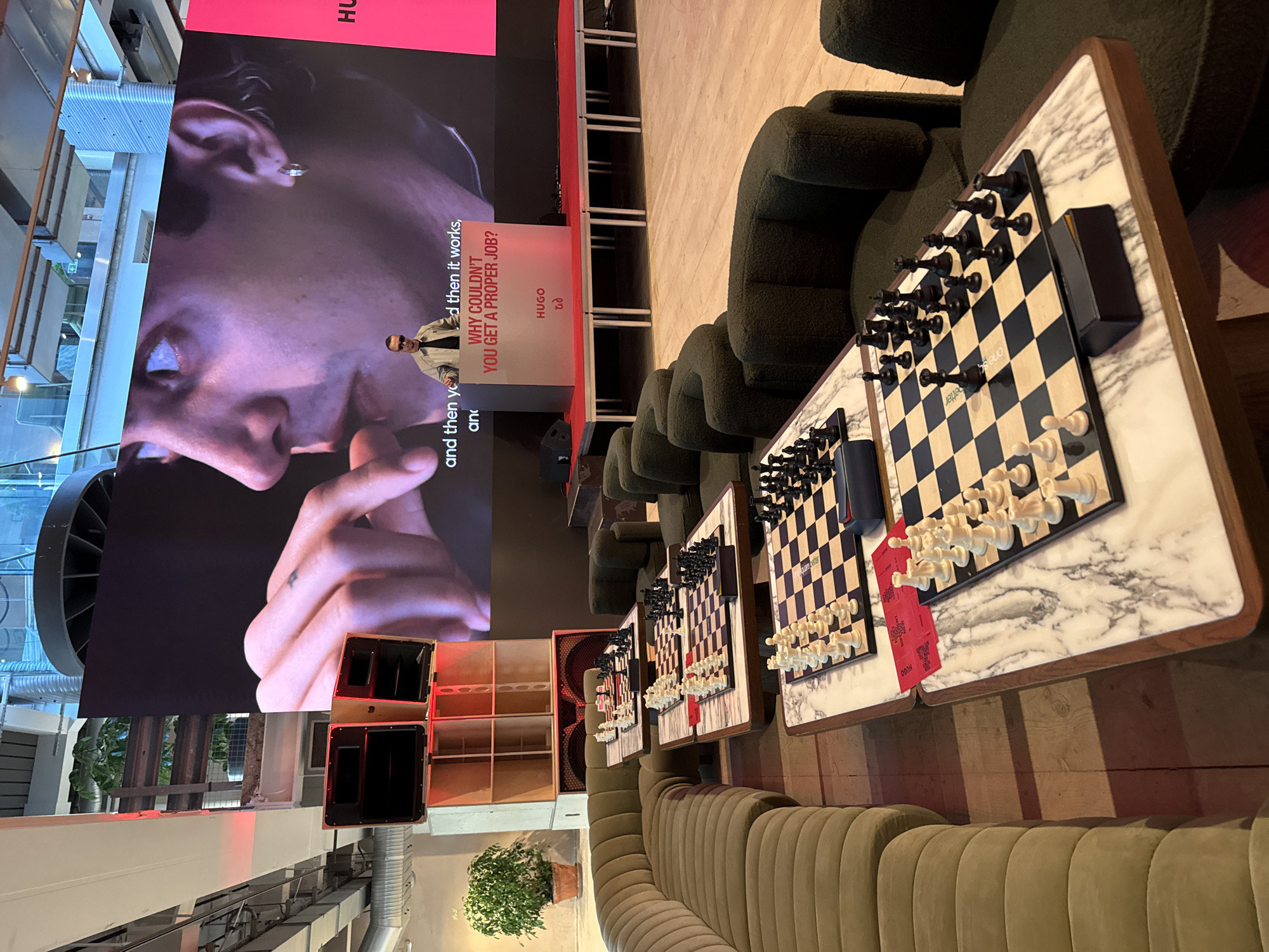 The photograph presents an interior scene in London, United Kingdom, showing an event or setup featuring chess boards on marble-topped tables, surrounded by dark green seating.
The foreground features three chess boards set up on marble-patterned tabletops. The chess pieces are set up in varying stages of a game or presentation. Beside one board, there's a stack of red cards or promotional material.
Behind the chess setups are rows of green, plush seating.
In the background, there's a large screen displaying a close-up image of a person's face. Adjacent to the face is promotional text associated with the HUGO brand. To the side of the screen, the architecture of what seems like a modern building is visible.
Additionally, there are light-colored wooden shelving units and a potted plant near the base of the screen. Speaker systems are also visible, placed near the screen. The composition indicates that this is likely an event space or lounge.
