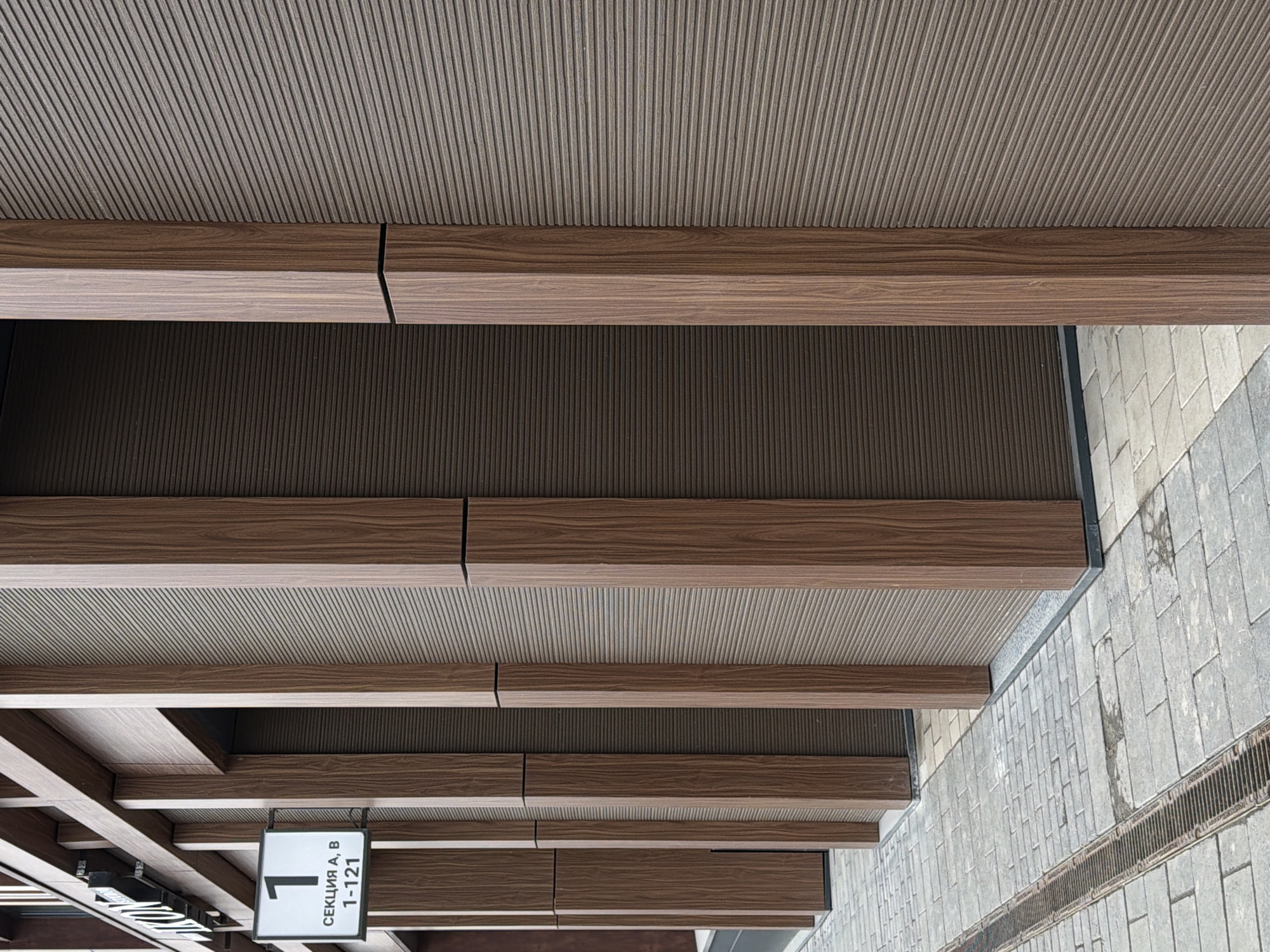 The image shows a portion of a building's exterior in what is likely Perm, Russia. The view is from below, looking up at a covered walkway or entrance area. The structure consists of a ceiling with a linear, textured surface, supported by wooden beams. There are light fixtures and signage hanging from the structure. The signage is partially visible and appears to have the number "1-121" on it and the word "СЕКЦИЯ". To the right of the structure is a paved area, likely a sidewalk, made of rectangular stones. There is a drain visible in the sidewalk. The scene appears to be outdoors during daylight hours.