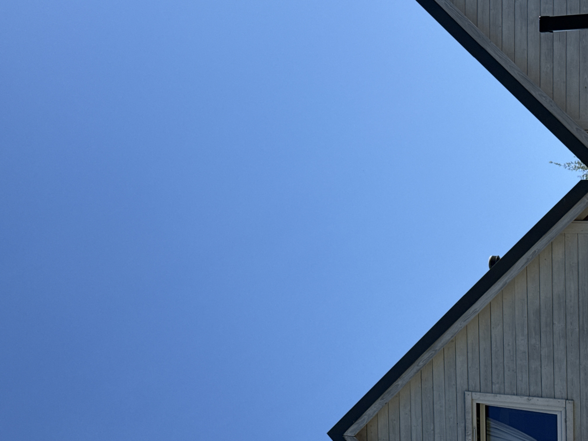 The image shows a section of two gabled roofs of houses against a clear blue sky. The roofs are angled, meeting in the center of the frame. The house on the left has a window with white curtains. The houses appear to be made of wooden planks, painted a light grey color. The rooflines are bordered with black trim. The environment appears to be outdoor, possibly in a residential area, with a clear sky suggesting fair weather. The location is identified as Ustronie Morskie, Poland. There are no people or animals visible in the image.