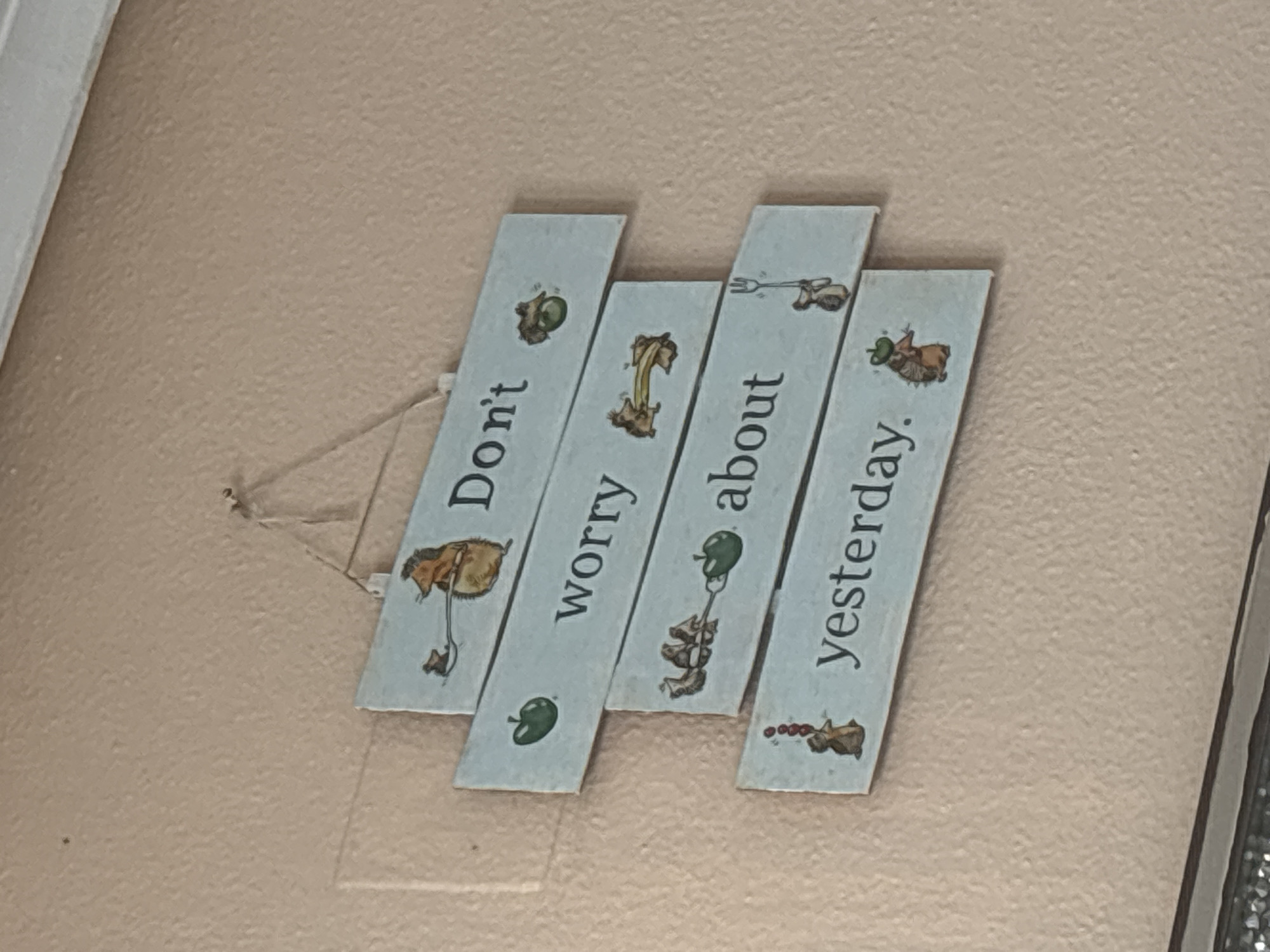 This is a close-up, eye-level, medium shot of a wall decoration. The decoration is made of four wooden slats arranged in a staggered pattern, tilted diagonally. Each slat has a phrase written on it in black, vertical lettering, with an illustration on each. The background is a textured, pale pink wall.
The text on the slats reads, from left to right: "Don't," "worry," "about," and "yesterday." Each slat also features a small, whimsical illustration. The first slat has a figure with large ears holding a leaf. The second slat depicts a brown, furry creature with a long tail on a yellow, arched object, possibly a bridge or rainbow. The third slat shows a green apple on a string, and the fourth slat has a small brown creature with red berries.
The overall tone of the decoration is cheerful and optimistic, suggesting a message of living in the present. The lighting appears to be from an indoor source, casting no strong shadows. There are no indicators of the time of day, weather, or specific location other than the provided context of Orihuela, Spain. The wall appears to be indoors, possibly in a home or public space.