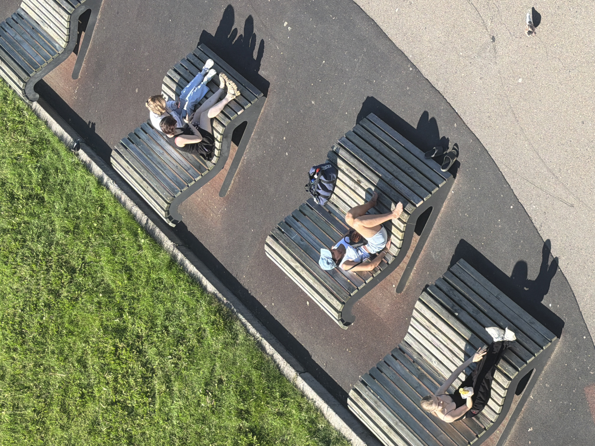 This is an aerial, high-angle, full shot of a park area with several modern, curved, wooden lounge benches arranged on a dark paved surface. To the left of the benches, a vibrant green lawn slopes upwards.
In the foreground, a young woman with blonde hair, wearing a black spaghetti strap top and black pants, is seated on the leftmost bench, holding a yellow object, possibly food, in her right hand and wearing white sneakers.
Further up and to the right, another young woman with short dark hair is seated on a bench, wearing a black dress. Next to her, a person with blonde hair, wearing a white top and blue jeans, is also seated on the same bench, looking at a phone. A dark blue duffel bag rests on the bench between them.
On the bench further up and to the right, a person wearing a white tank top and denim shorts is reclining, with a blue cap on their head and a dark bag on their lap. Their legs are bent.
In the background, another bench is partially visible, empty. The scene is illuminated by bright sunlight, casting distinct shadows. The paved surface appears to be a walkway or park area. The specific location within Krasnoyarsk, Russia, is not identifiable from the visual information.