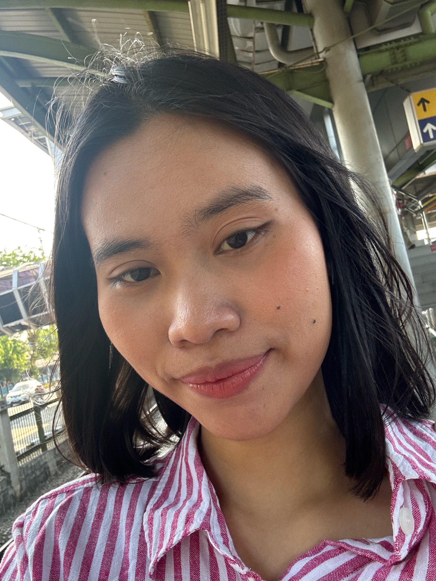A selfie taken from a slightly low angle features a young East Asian woman with shoulder-length, dark hair. She is wearing a pink and white striped button-up shirt. The background suggests an outdoor urban environment, possibly a train station or elevated walkway, with visible infrastructure like poles, signage with arrows indicating directions, and a glimpse of a street with cars and foliage below. The lighting indicates it is daytime.