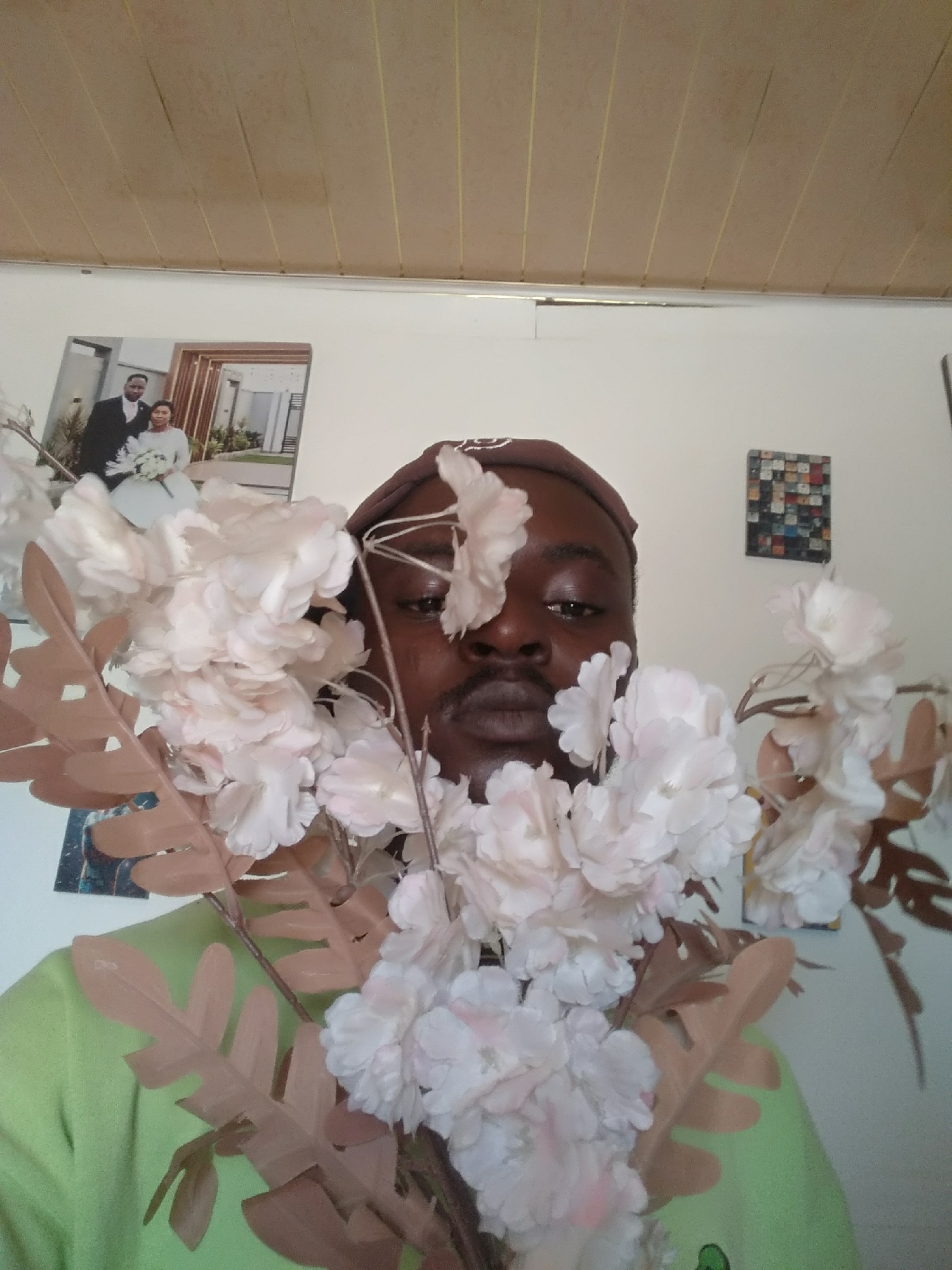 A dark-skinned man is captured in a medium close-up, largely obscured by a large bouquet of artificial flowers. He is positioned centrally, looking directly at the viewer with a neutral expression. Only his eyes, nose, and mouth with a mustache are visible through the light pink and white petals and brownish-pink leaves of the floral arrangement. He is wearing a light green shirt and a brown head covering, possibly a cap or bandana.
The scene is indoors, with light-colored walls in the background. On the left side, a framed wedding photograph hangs on the wall, showing a man in a black suit and a woman in a white wedding dress holding a bouquet. On the right wall, a smaller, square, framed artwork or collage, made of various small, colorful rectangular pieces, is visible. The ceiling is paneled, with light brown rectangular sections separated by thin yellow lines. The overall lighting appears bright and even, suggesting an indoor setting with ample light. There is no visible text in the image.