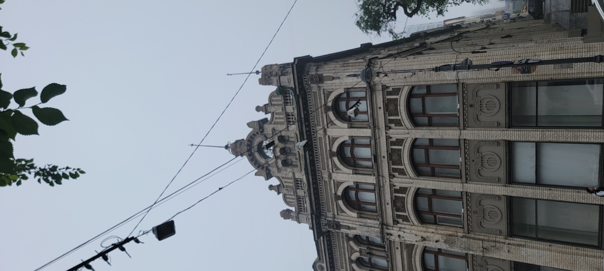 The image shows a multi-story building with ornate architectural details, possibly in an older part of Vladivostok, Russia, on an overcast day. The building's facade is constructed of light-colored bricks and features arched windows with dark frames. Decorative elements such as pilasters, cornices, and sculpted panels are visible. Electrical wires and a street lamp are present in the foreground. A single person is standing near the bottom left of the frame, partially obscured. The sky is a uniform gray, suggesting cloudy weather. Trees are visible on the right side of the image, and some branches with leaves hang into the top portion of the frame. The overall impression is of a historical urban setting.