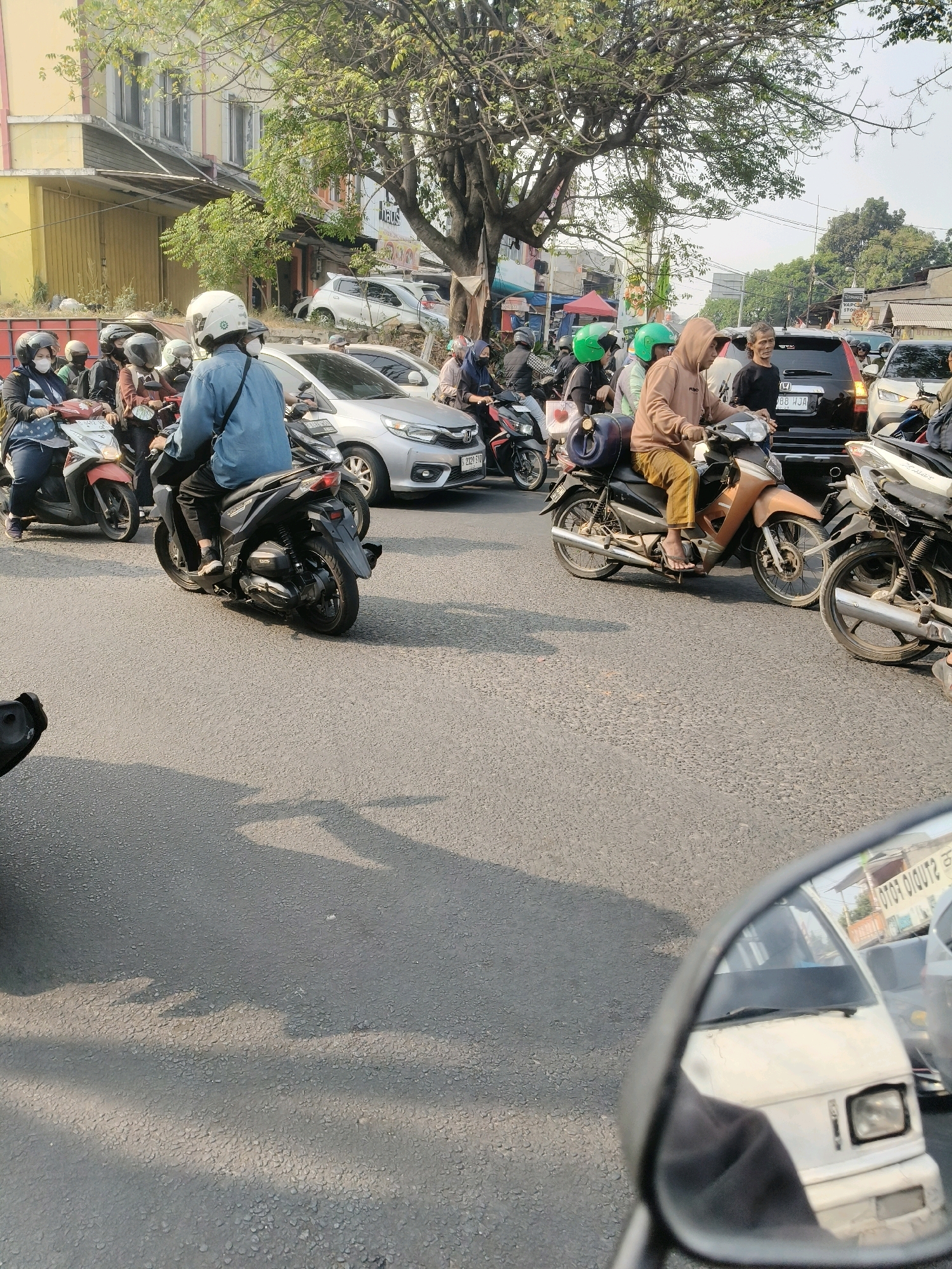 The image depicts a traffic scene on a paved road in South Tangerang, Indonesia, likely during daylight hours. Numerous motorcycles, many with riders wearing helmets, are present, indicating a typical urban environment with significant motorcycle usage. One individual on a motorcycle is wearing a blue long-sleeved shirt and black pants, with a white helmet. Another rider in the foreground is wearing a light brown hooded jacket and brown pants, also on a motorcycle. Several cars are visible in the background, suggesting a busy road. Buildings line the sides of the street, with one building on the left featuring yellow and grey facades. Trees with green foliage are also present, casting shadows on the road. The scene is captured from a low angle, possibly from another motorcycle, with a rear-view mirror in the bottom right corner reflecting a white vehicle and signage.