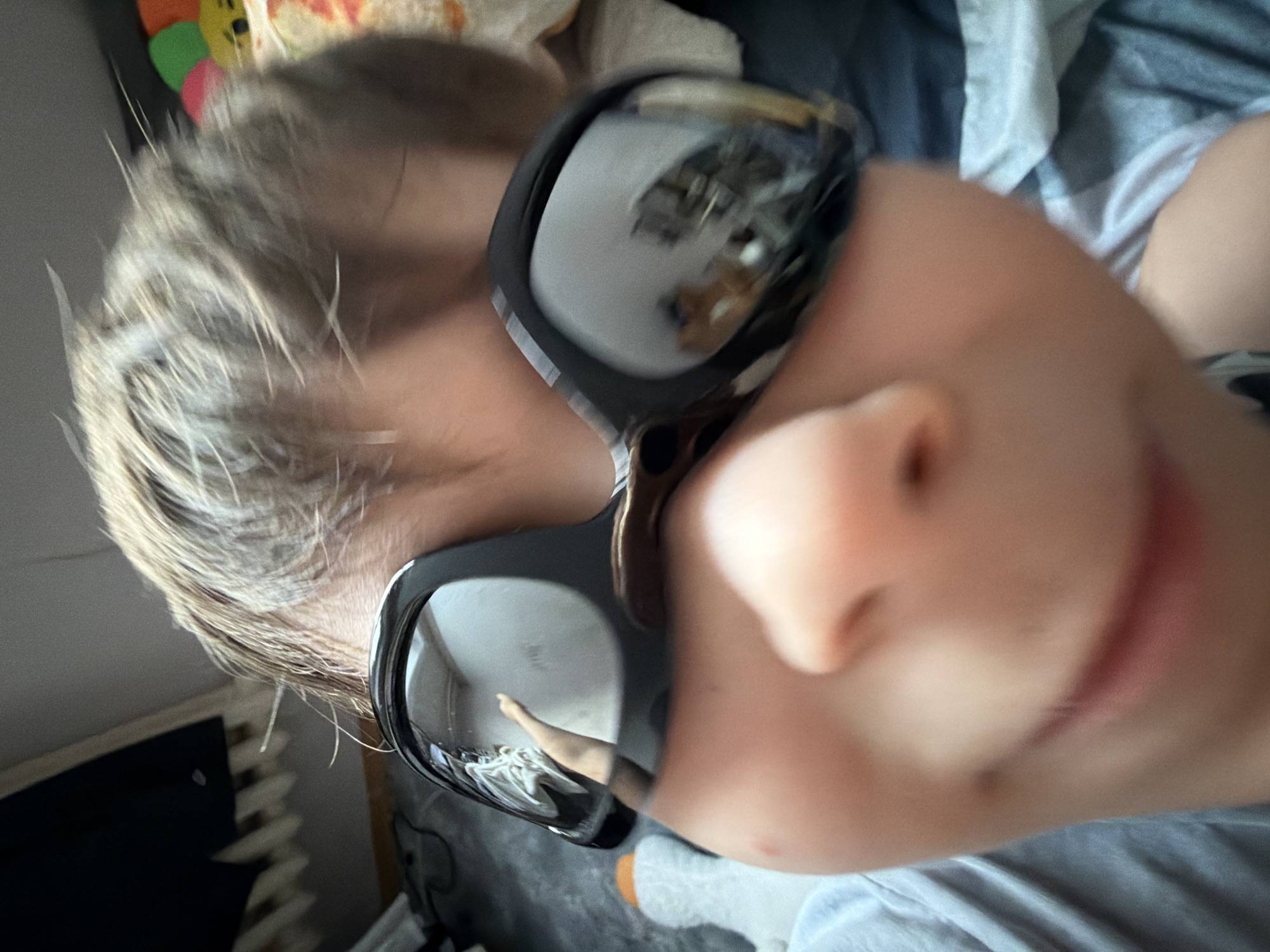 A child is lying down, facing the camera, wearing black sunglasses. The reflection in the sunglasses shows a room interior. The child has light blonde hair and is wearing a light blue shirt. A patterned blanket or toy is visible in the upper left corner. A radiator can be seen in the lower left corner. The photo appears to be taken from a close-up, low-angle perspective. It is not possible to identify the location as Perm, Russia or any other city based on the visual information provided.