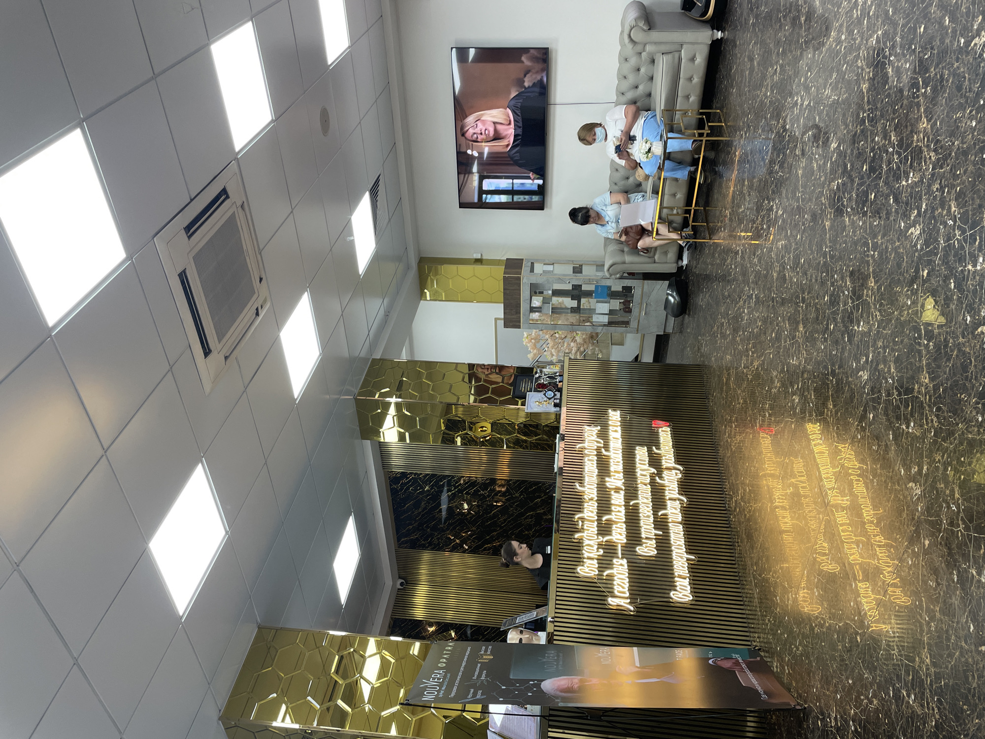 The image depicts the interior of a modern beauty salon in Almaty, Kazakhstan. The reception area is characterized by opulent gold accents, including hexagonal patterned pillars and vertical ribbed paneling. A neon sign with Cyrillic text is prominently displayed on the reception desk, reflecting onto the polished marble floor.
In the background, a woman in a black uniform stands behind the reception desk, appearing to be an employee. To the right of the reception area, a gray tufted sofa is occupied by two women. One woman, wearing a light blue dress and glasses, is looking at a document, while the other, in a white t-shirt and blue pants, is using her mobile phone. A small glass and gold coffee table sits in front of them.
A large flat-screen television mounted on the wall displays a woman speaking. The ceiling is equipped with recessed fluorescent lighting panels and an air conditioning unit. A display case with beauty products is visible to the left of the sofa. A promotional banner for "NOUVERA" is positioned near the reception desk. The overall ambiance suggests a high-end establishment.