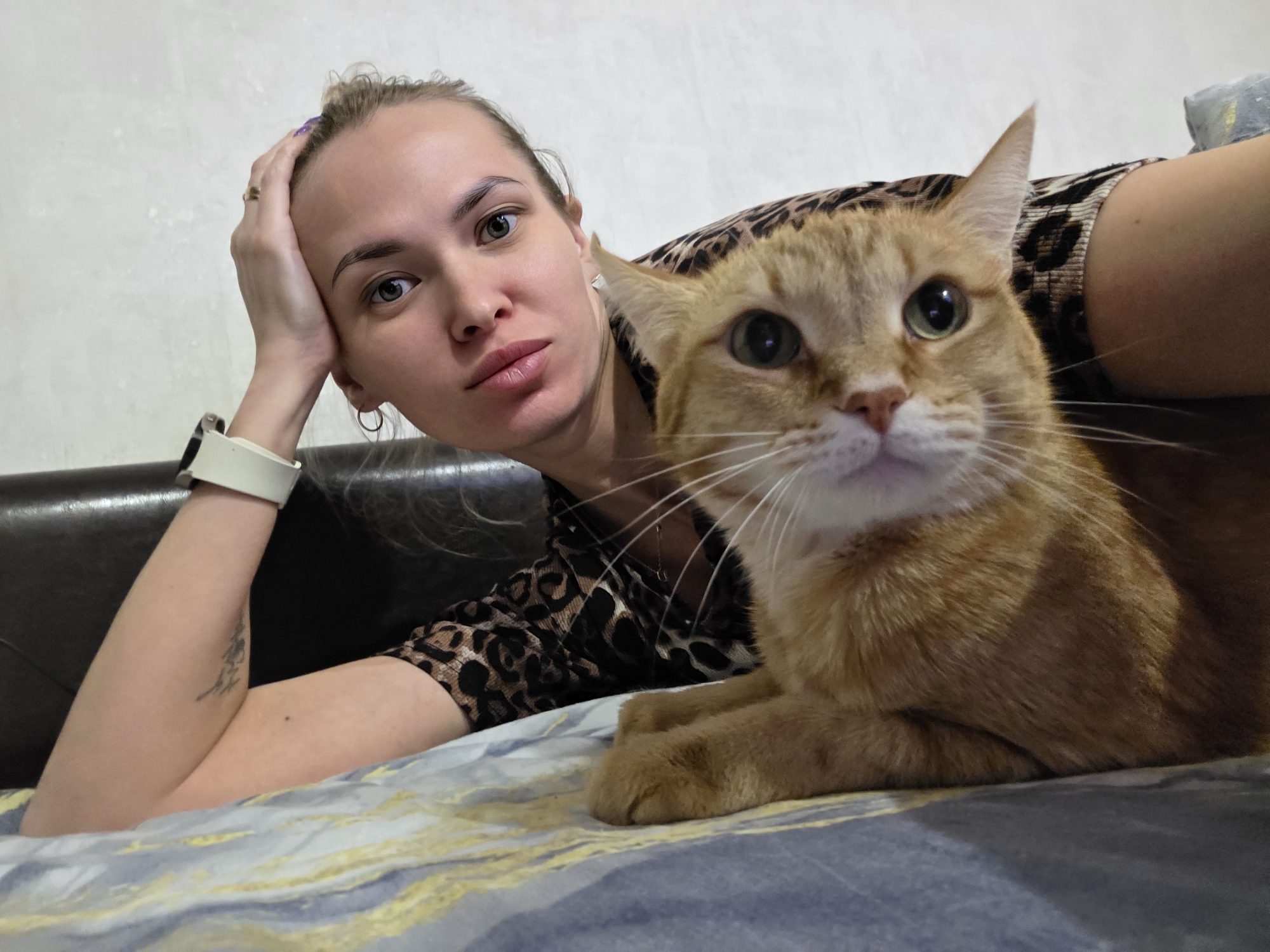 A woman and an orange cat are lying on a bed. The woman is on her left side, propped up on her left arm, looking at the camera. She has blonde hair pulled back in a bun, light skin, and is wearing a leopard print top. She has a smartwatch on her left wrist and a small tattoo on her forearm. The cat is in the foreground, facing forward, with its paws resting on the bed. The bed has a blue and gray patterned comforter. A black leather headboard is visible in the background. The location is Chaikovsky, Russia.