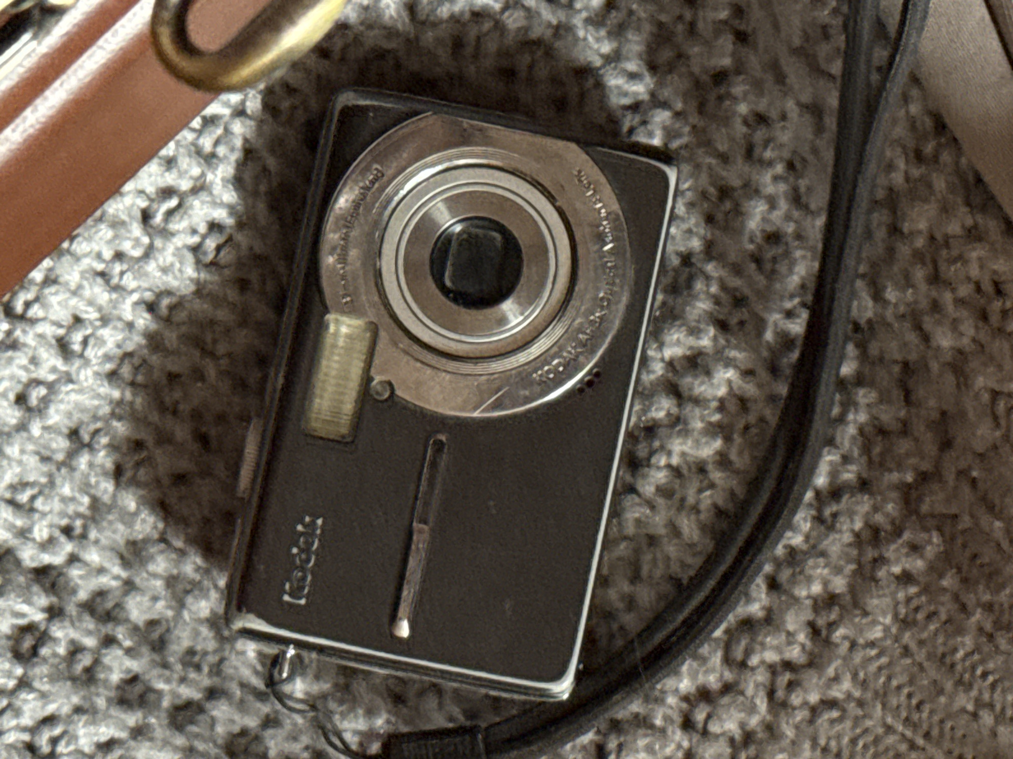 This media file features a close-up, top-down view of a black Kodak digital camera resting on a grey, textured surface, likely a carpet or rug. The camera is positioned diagonally in the frame, with the lens and controls facing upwards.
The most prominent subject is the camera itself. It is a compact digital camera with a silver-toned lens assembly encircled by concentric rings. Text on the lens assembly is partially visible and appears to read "KODAK" and possibly model-specific information like "DIGITAL" and "ZOOM". Below the lens, the "Kodak" logo is clearly visible in a silver, stylized font. A small, rectangular flash or LED indicator is located to the left of the lens. A thin, silver metallic strip, possibly a button or indicator, is centered on the camera's body. A black strap is attached to the bottom left of the camera and trails off the edge of the frame.
To the upper left of the camera, a portion of a brown leather strap with a brass-colored clasp is visible, suggesting the camera might be part of a bag or attached to a larger accessory. The background is a dense, textured grey surface with visible fibers, indicative of a domestic indoor setting.
There is no indication of people, specific weather conditions, or time of day. The setting is likely indoors, given the presence of carpet and the absence of natural light cues. The overall mood is neutral, focusing on the object. The visible text identifies the brand of the camera as "Kodak". No specific location cues for Izhevsk, Russia are evident within the image itself.