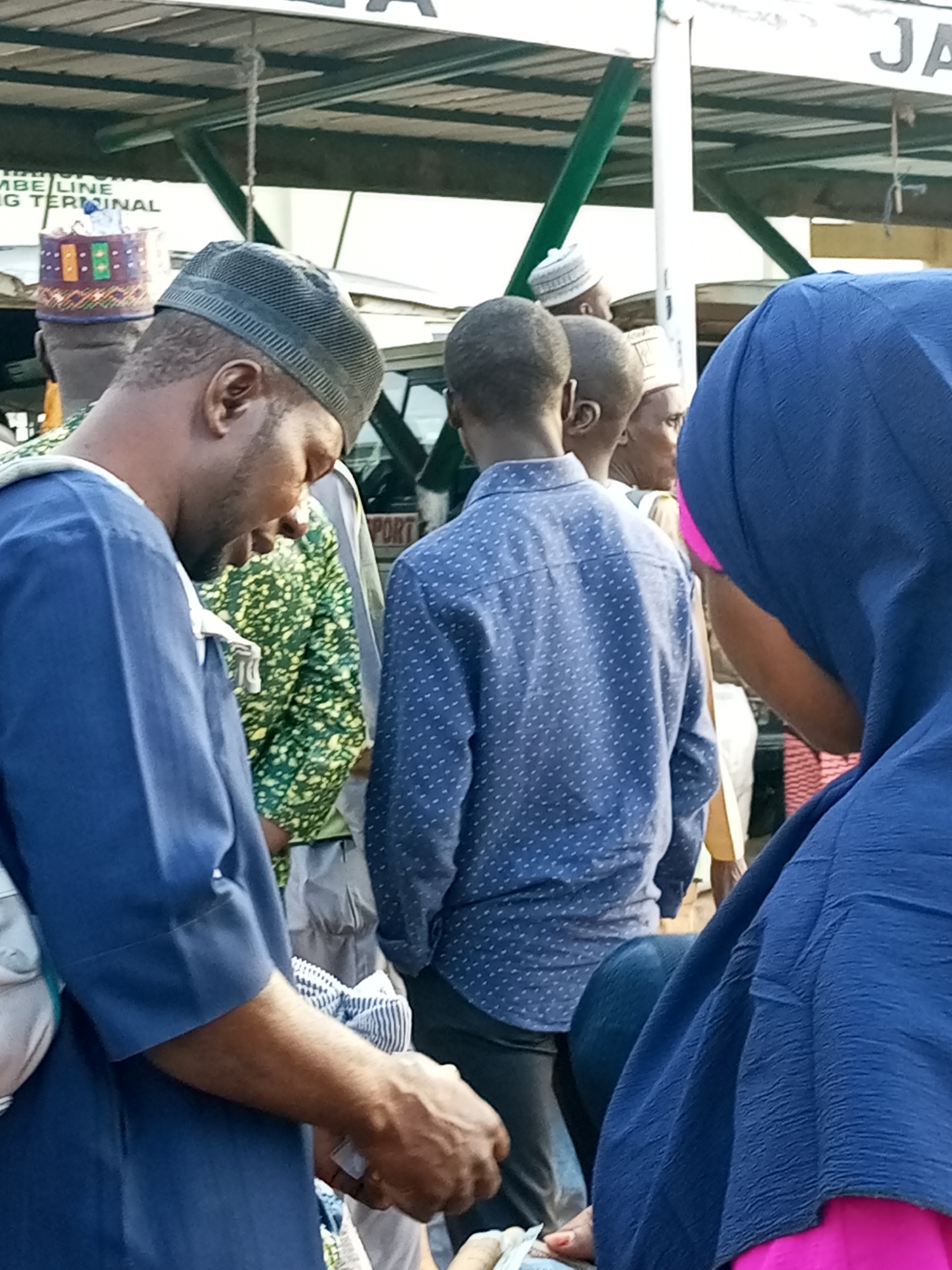 The image depicts a gathering of people at the Gombe Central Market in Gombe, Nigeria. In the foreground, a man wearing a blue tunic and a grey cap is facing down and appears to be engaged in a transaction, holding what looks like cash in his hand. To his right, another man in a blue patterned shirt is visible. Behind them, several other individuals are present, some wearing traditional attire and caps. The background reveals market stalls and a sign that partially reads "Gombe Line Shipping Terminal". The overall scene suggests a busy market environment with people interacting and possibly buying or selling goods.