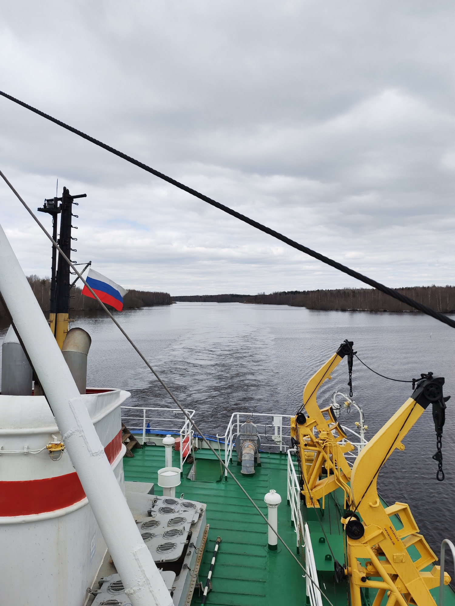 The image captures a view from the stern of a ship traveling along a wide river or canal, likely in Uzhla, Russia. The sky is overcast with a uniform layer of gray clouds, indicating a cloudy day. Both banks of the waterway are densely forested with bare deciduous trees, suggesting the season is late autumn or early spring.
In the foreground, elements of the ship's structure are visible. The deck is painted a practical green. On the left, a portion of the ship's white hull, accented with a red horizontal stripe, and a thick white mast or support pole are prominent. Further left, a darker mast-like structure displays a Russian flag, horizontally striped in white, blue, and red, fluttering in the breeze.
The right side of the deck is dominated by two robust, bright yellow crane-like structures, possibly davits, equipped with chains and hooks, designed for launching or retrieving objects. Visible text on one of these yellow components reads "ИСПОЛ.022-L" and "ТПРЭ.02.26", likely identifying markings or specifications. Other deck features include white railings, various metallic hatches with circular closures, and a white ventilation pipe.
The ship's movement is evident from the distinct wake trailing behind it in the dark water. Several thick, black ropes or cables belonging to the ship's rigging stretch diagonally across the upper portion of the frame. The river extends into the distance, bordered by the unbroken treeline on either side, conveying a sense of progression through a natural, inland waterway.