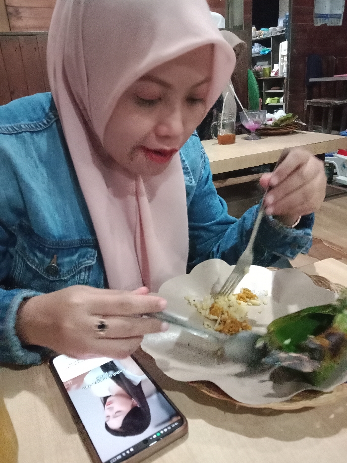 An adult female is depicted seated at a wooden table within an eatery located in Kebonsari, Indonesia. She is wearing a light pink hijab and a blue denim jacket. Her right hand holds a metal fork, positioned to gather food from a serving of white rice and a yellow-orange granular topping presented on a fluted paper plate, which rests on a woven tray. Her left hand holds a metal spoon. Adjacent to her plate is a portion of food wrapped in a green leaf. Her mouth is slightly open. A smartphone is placed face-up on the table to her left, displaying an image of another female. In the background, a light-colored wooden table holds a glass mug with a straw containing a brown liquid, and a clear glass with a pink substance and a straw. Wooden walls, shelves with assorted items, and additional wooden seating are visible in the dimly lit environment.