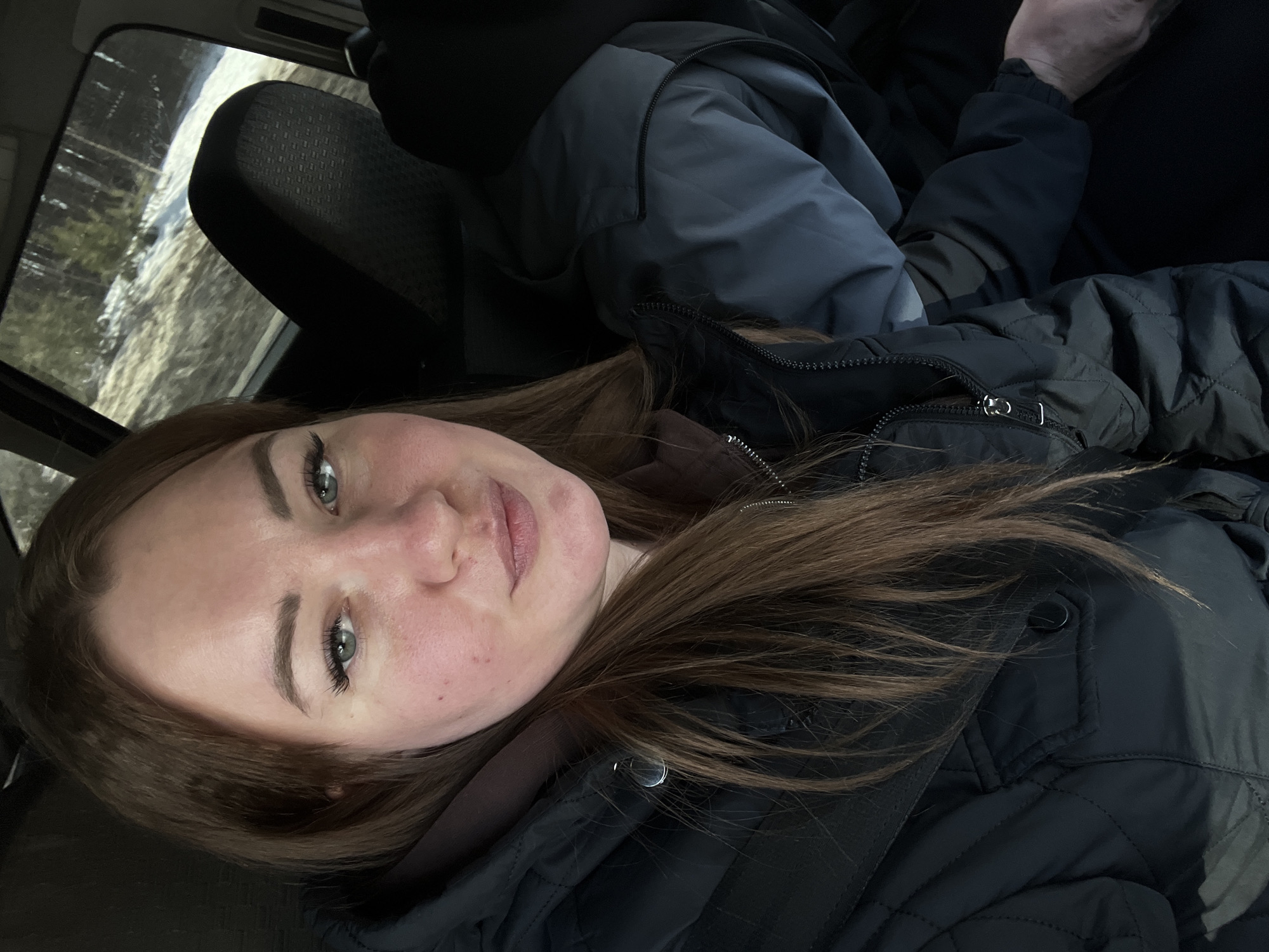 A selfie taken inside a car. The person is wearing a black quilted jacket and a seatbelt. The background shows a blurry landscape through the car window, suggesting a rural or suburban area. Without more precise details, providing a location in the city of Chaikovsky, Russia is not possible.