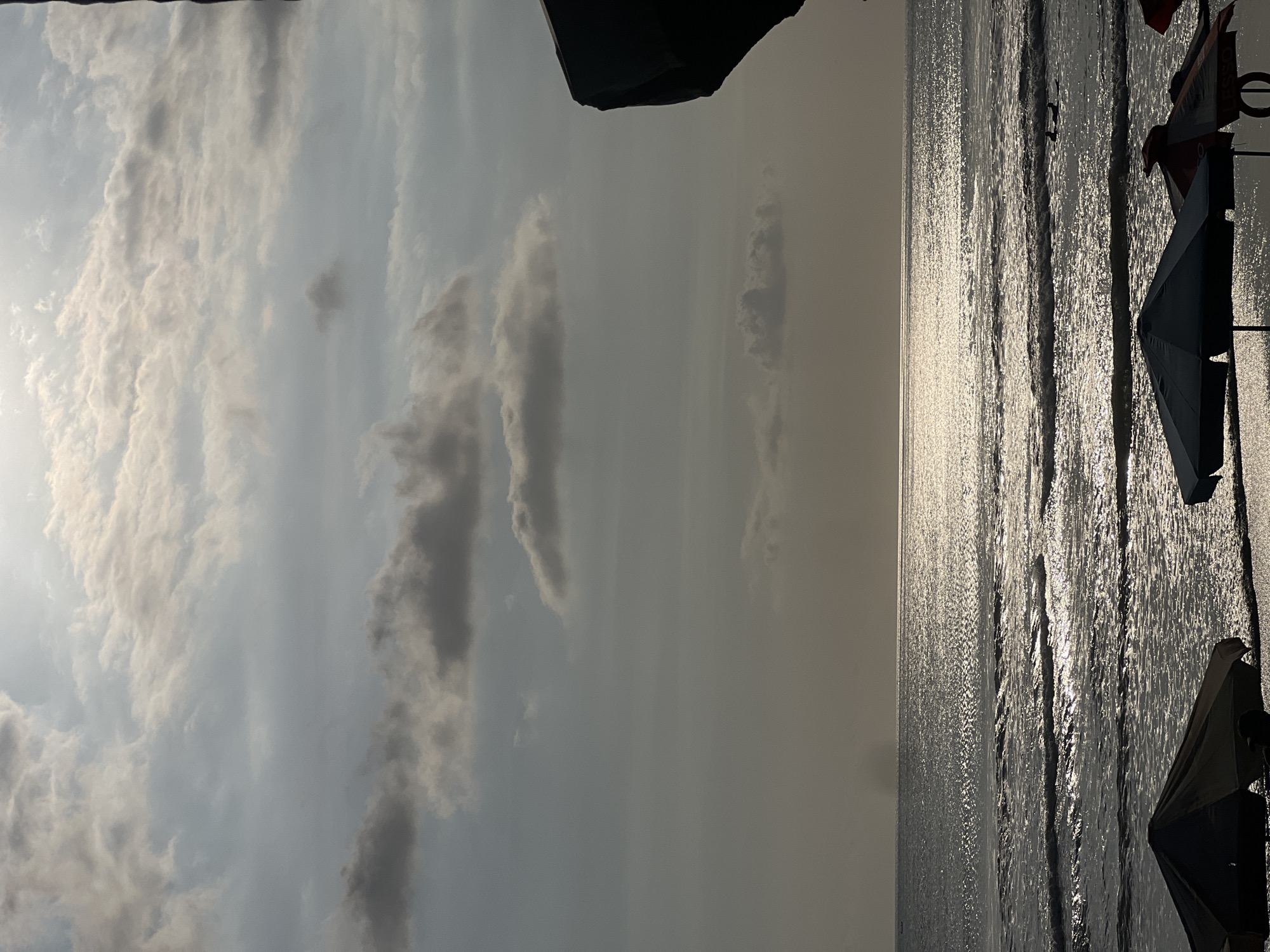 The image depicts a serene beach scene in Kuta, Indonesia, during what appears to be either sunrise or sunset, given the soft, diffused light and the way the sun's rays reflect off the ocean. In the foreground, beach umbrellas are set up on the sand, casting shadows and providing a sense of a recreational area. The ocean stretches out to the horizon, with gentle waves breaking on the shore, their surfaces shimmering with reflected light. The sky above is filled with a mixture of light and dark clouds, adding a dramatic and textured element to the scene. A faint silhouette of what could be a distant boat or landmass is visible on the horizon. The overall mood of the image is tranquil and atmospheric, capturing the natural beauty of the Kuta coastline.