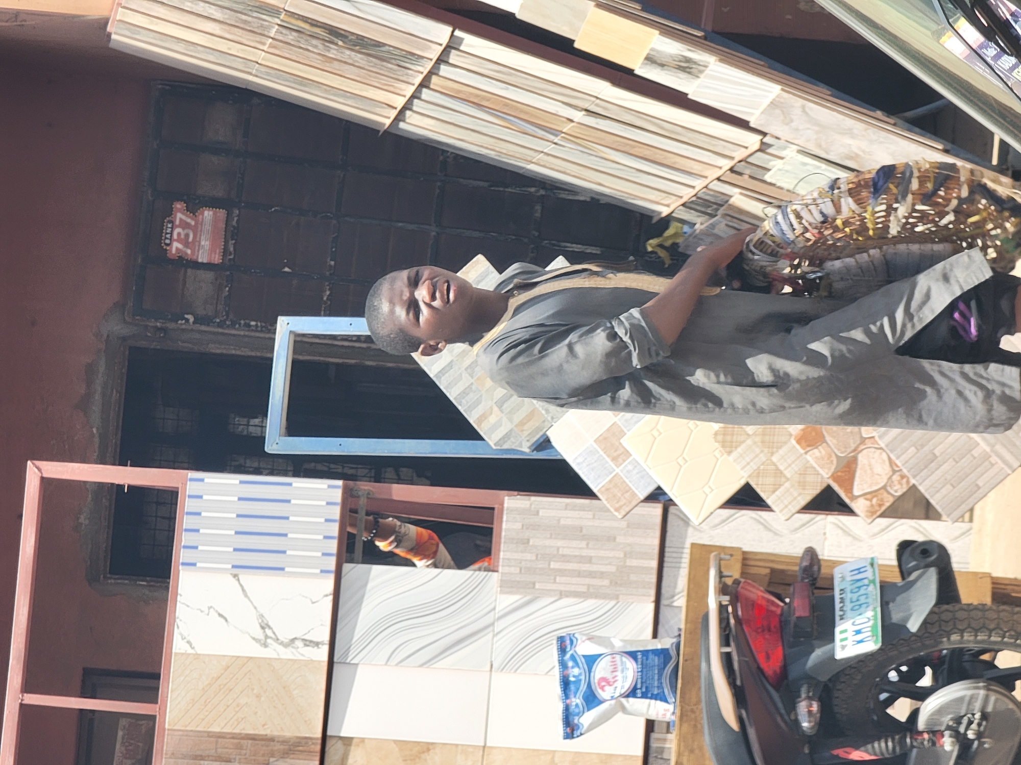 The image, taken in bright daylight and rotated clockwise, captures a young man standing amidst a display of ceramic tiles, likely in a market or shop setting in Maiburiji, Nigeria. The central figure, a young male, wears a dark grey long-sleeved tunic with light brown or gold trim and holds a woven basket or container. He looks towards the viewer with a neutral expression.
The surrounding environment is filled with various types of tiles, including those mimicking wood, marble, and patterned designs, stacked and arranged for display. Behind the man is a reddish-brown building with a window protected by dark metal grilles. A red sticker is visible on the window grille, bearing the text "BANK" and the numbers "737", indicative of a mobile banking service often used in Nigeria.
In the lower right, the rear section of a motorcycle or scooter is visible, featuring a green and white license plate that clearly shows "KANO" and "KMC 959YH," confirming the location in Kano State, Nigeria. On the ground, a white and blue bag with "R White" printed on it can be seen, possibly containing building materials. Another person's arm, wearing an orange and white patterned sleeve, is visible through an opening in the background. The scene depicts an active commercial area under clear skies.