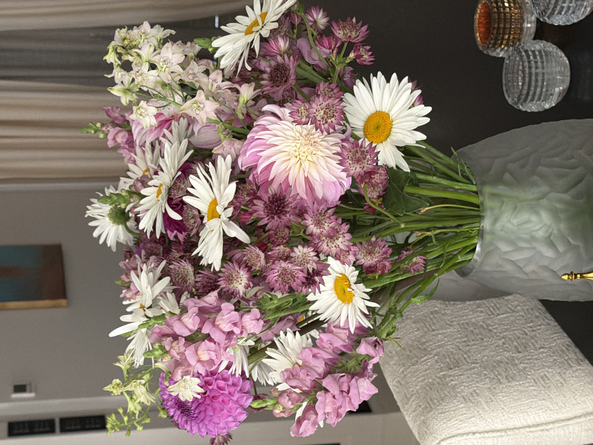 A bouquet of flowers, predominantly pink and white daisies with purple accents, is arranged in a frosted glass vase. The vase sits on a dark surface, possibly a table, beside a textured white chair. In the background, sheer curtains hang, and a framed artwork is visible on a grey wall. The scene appears to be indoors, likely in a residential setting, and there is no indication of it being located in Moscow, Russia.