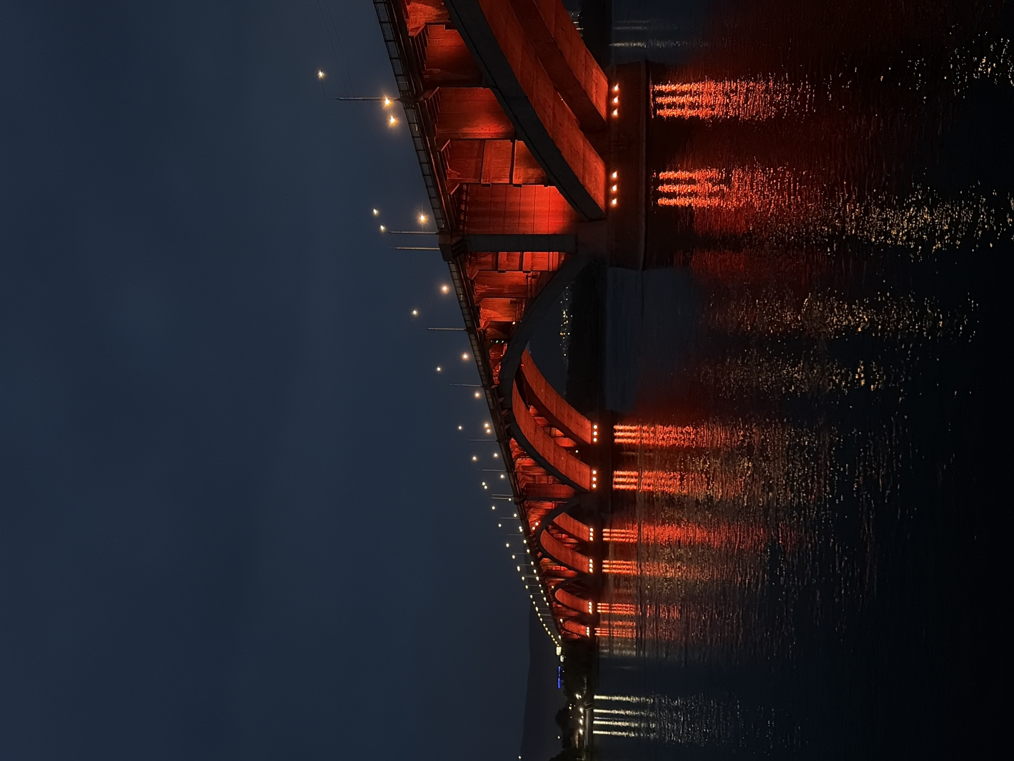 The image depicts the "Kommunalny Bridge" in Krasnoyarsk, Russia, at night. The bridge, illuminated by vibrant red lights on its arches and supports, spans across the Yenisei River. The lights cast strong reflections on the dark, rippling water. The sky above is a deep, dusky blue, with a few distant lights from the city visible on the horizon. The bridge itself is made of concrete and features multiple arched spans supported by massive piers. Streetlights are also visible along the top of the bridge. There are no people or vehicles clearly visible in the image.
