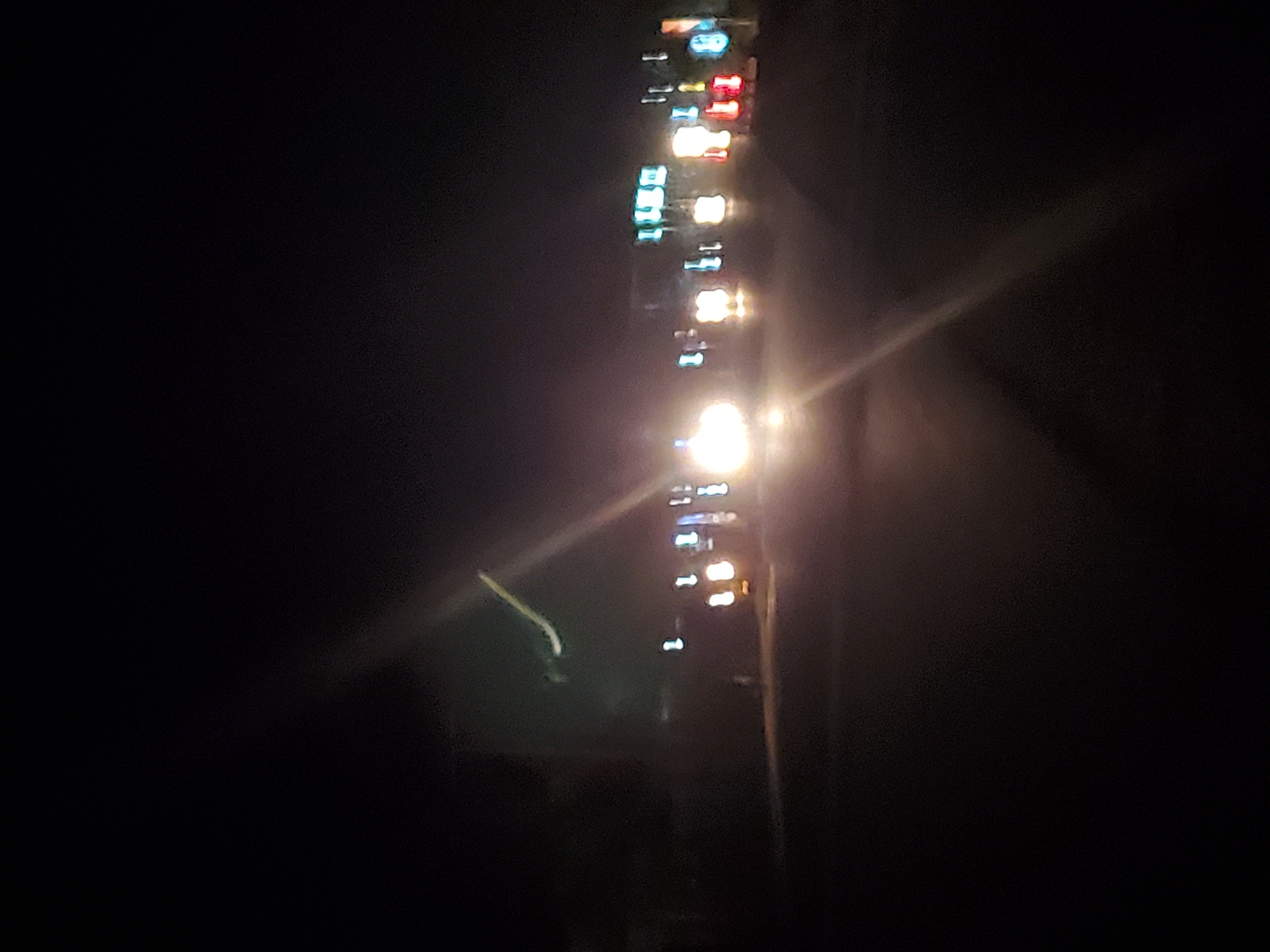 This is a nighttime photograph taken in Jalingo, Nigeria, depicting a street scene at night. The image is characterized by low light conditions and motion blur, suggesting it was taken from a moving vehicle. Several vehicles with their headlights illuminated are visible on the road, creating bright streaks of light. On the sides of the road, there are numerous blurred lights of various colors, indicative of businesses or advertisements. The overall environment appears to be a commercial or populated area within the city, with buildings and possibly streetlights contributing to the ambient illumination. The sky is dark, and there are no discernible individuals or specific activities captured in detail due to the photographic conditions.