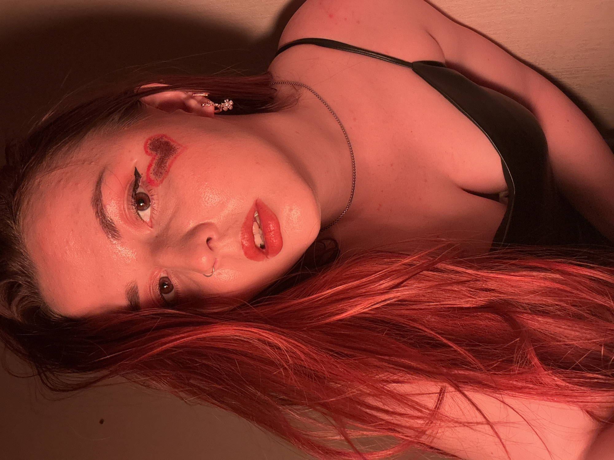 A woman with red hair and heavy makeup lies on her side. Her skin is fair, and she has a heart-shaped mark on her forehead near her left eyebrow. She wears a black tank top and a silver necklace. Her hair is spread around her face and upper body. The scene is lit with a red light. There is no evidence of location in Izhevsk, Russia.