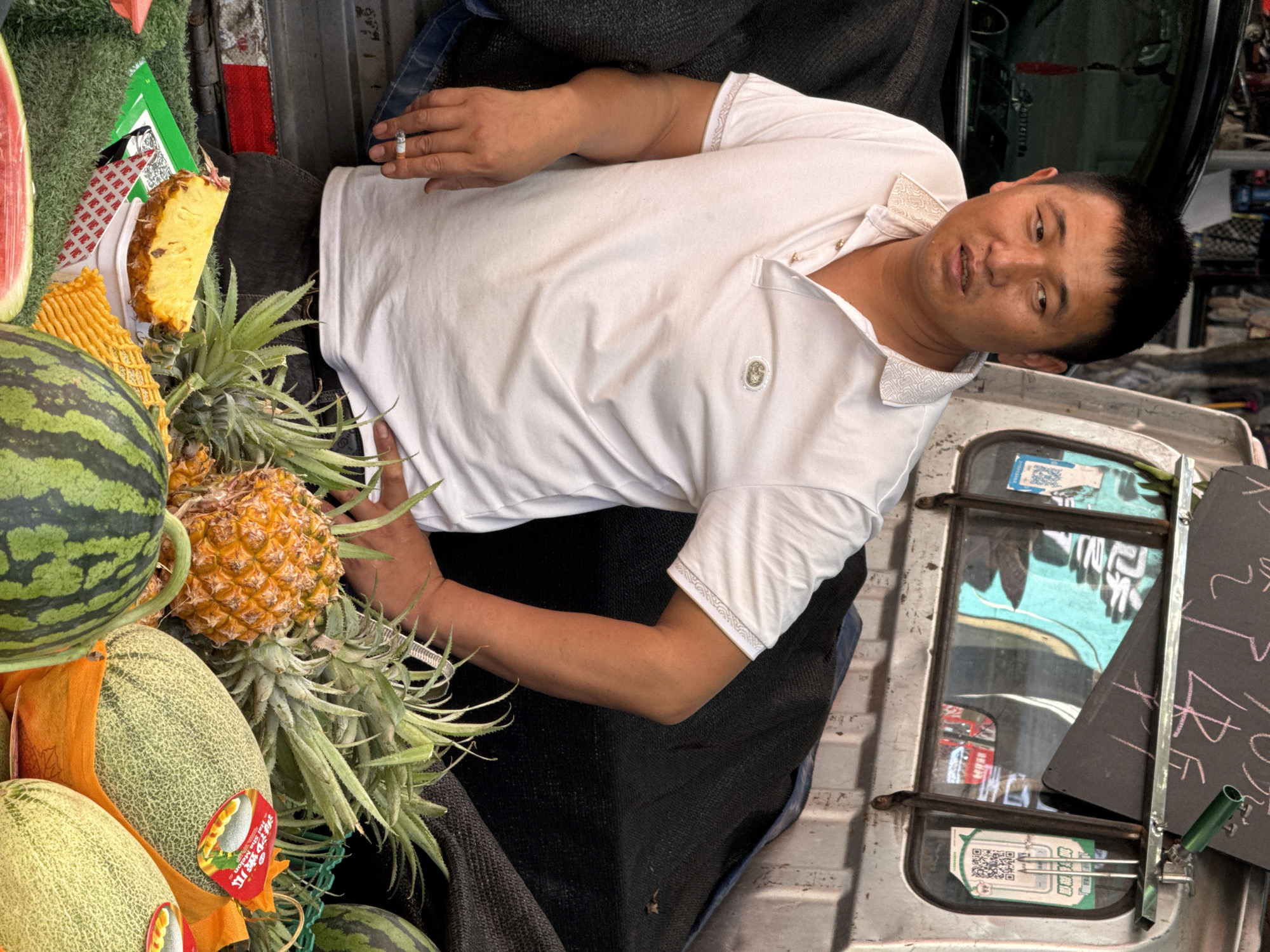 A middle-aged Asian man wearing a white polo shirt is holding a cigarette in his right hand. The man is surrounded by various fruits including watermelons, pineapples, and cantaloupes. He is leaning against the back of a silver-colored vehicle with a sign posted on the back. The window of the vehicle has a QR code sticker. The location is identified as the city of Beibei, China.