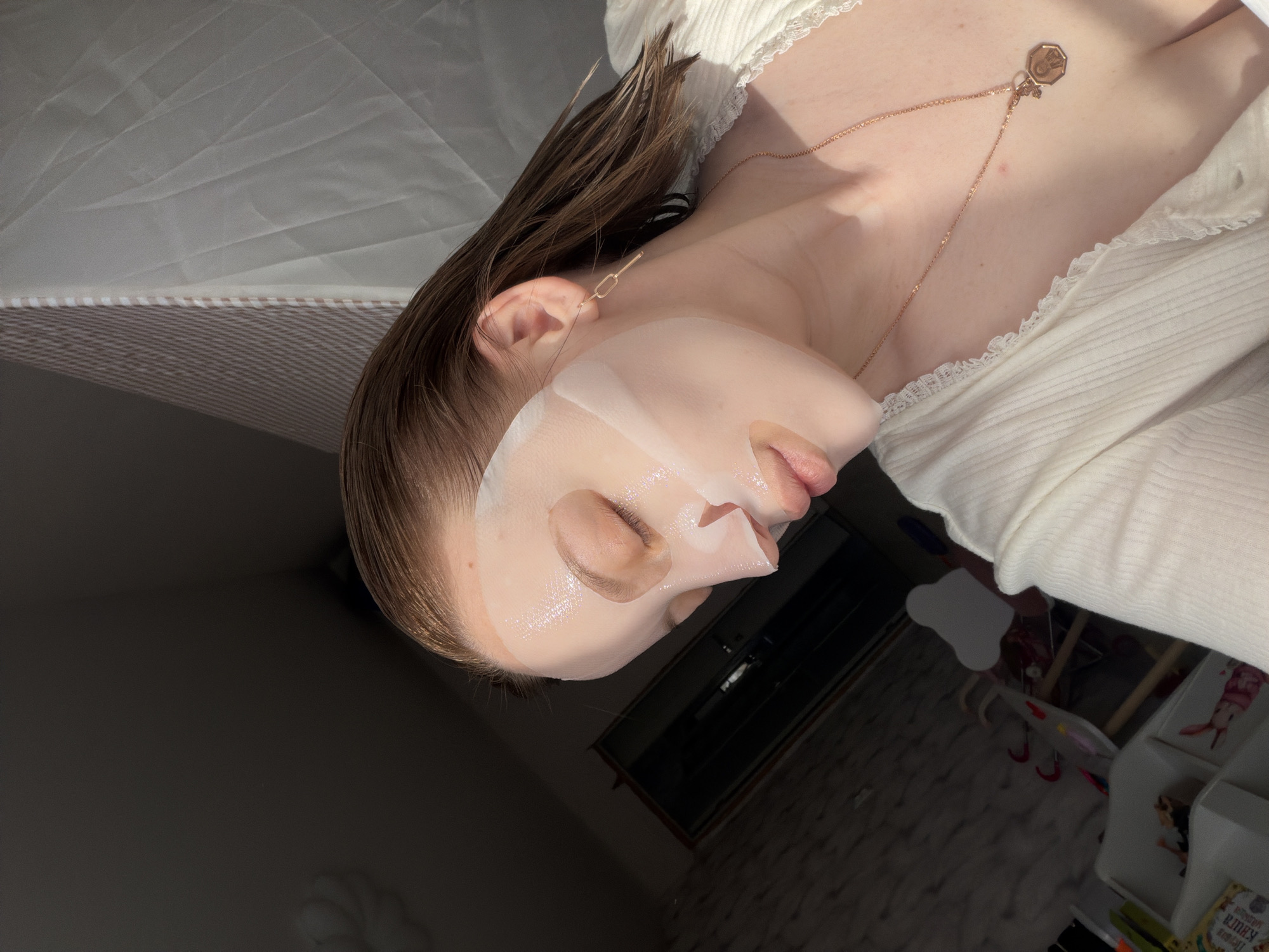 A young, light-skinned woman is lying on her back on a bed, with a white sheet mask applied to her face. She is wearing a white, lace-trimmed top and a gold necklace. Her hair is wet and slicked back. The room is dimly lit, with sunlight streaming in from the right side of the frame. In the background, there is a white bedside table with children's books and toys. There is no information available within the image to confirm the location of the scene. Therefore, the city of Reshetnikovo, Russia, is not discernible.