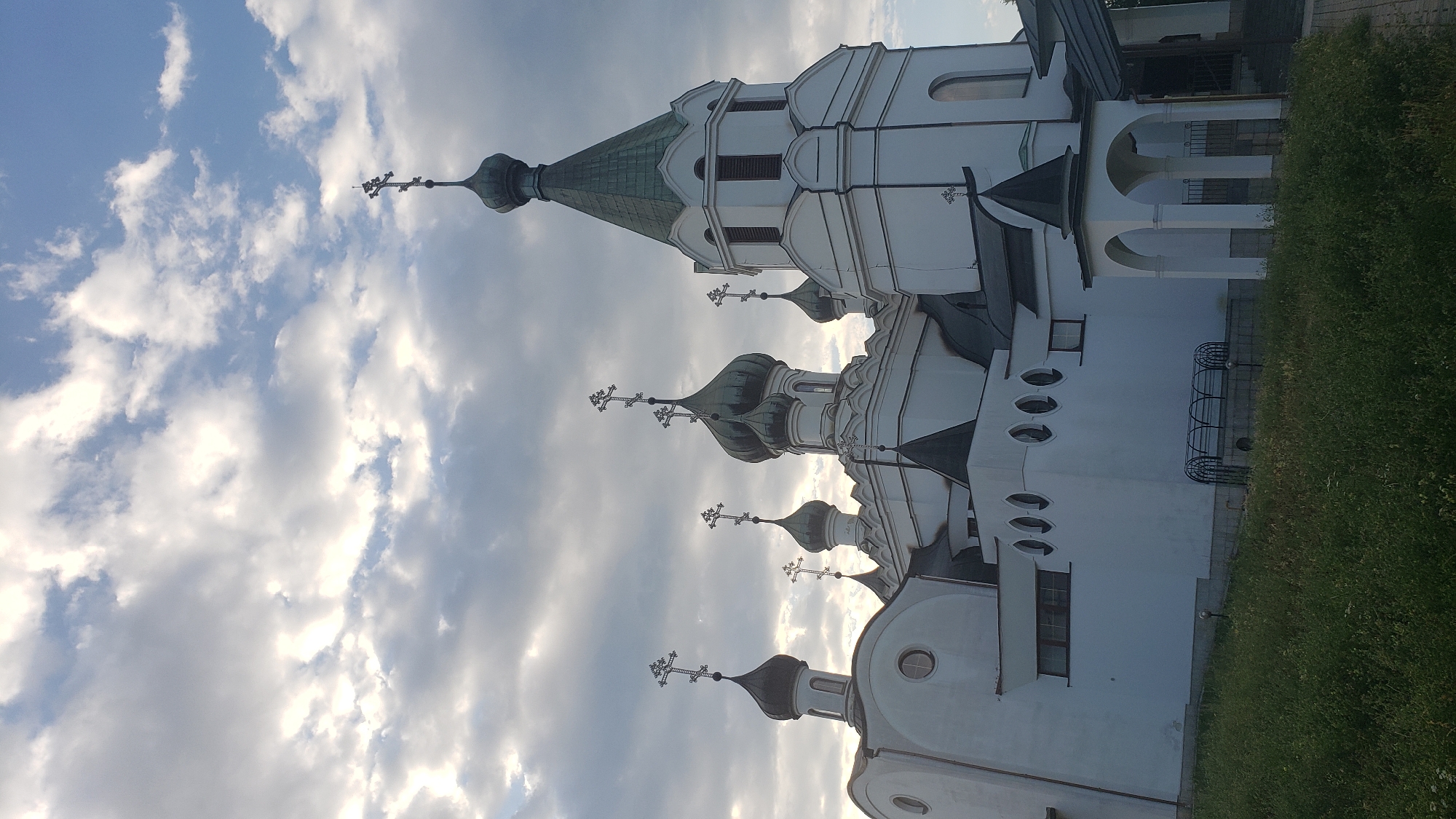 An exterior, low-angle shot captures the white facade of an Orthodox church against a dramatic sky filled with light and dark clouds. The church features multiple onion domes, each topped with a cross. The main structure is a complex arrangement of white walls, dark roofs, and arched entrances. In the foreground, green grass and foliage lead up to the church's grounds. There are no people visible in the image. The scene appears to be located in the city of Prešov, Slovakia, likely at the Orthodox Cathedral of Saint Alexander Nevsky, given its architectural style.
