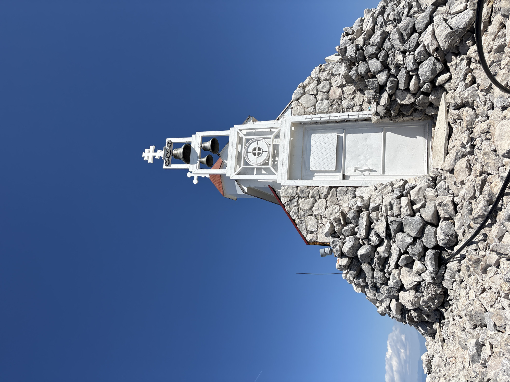 A low-angle, slightly rotated, outdoor shot depicts a small, white building constructed of stone and painted metal components, set against a clear, bright blue sky. The building is situated atop a rock formation. At the far left of the building is a white, metal structure supporting three small bells and topped with a Christian cross. To the right of the bells is a white, rectangular structure featuring a circular design. Adjacent to this is a square, metal panel with a diamond-patterned surface. To the right of the metal panel are two white, metal doors set into the stone wall of the building. A small portion of a red roof is visible on the side of the building. An electrical cable runs along the top right of the stone structure. The location is deduced to be Mali Mikulići, Montenegro, based on the architectural style and landscape.