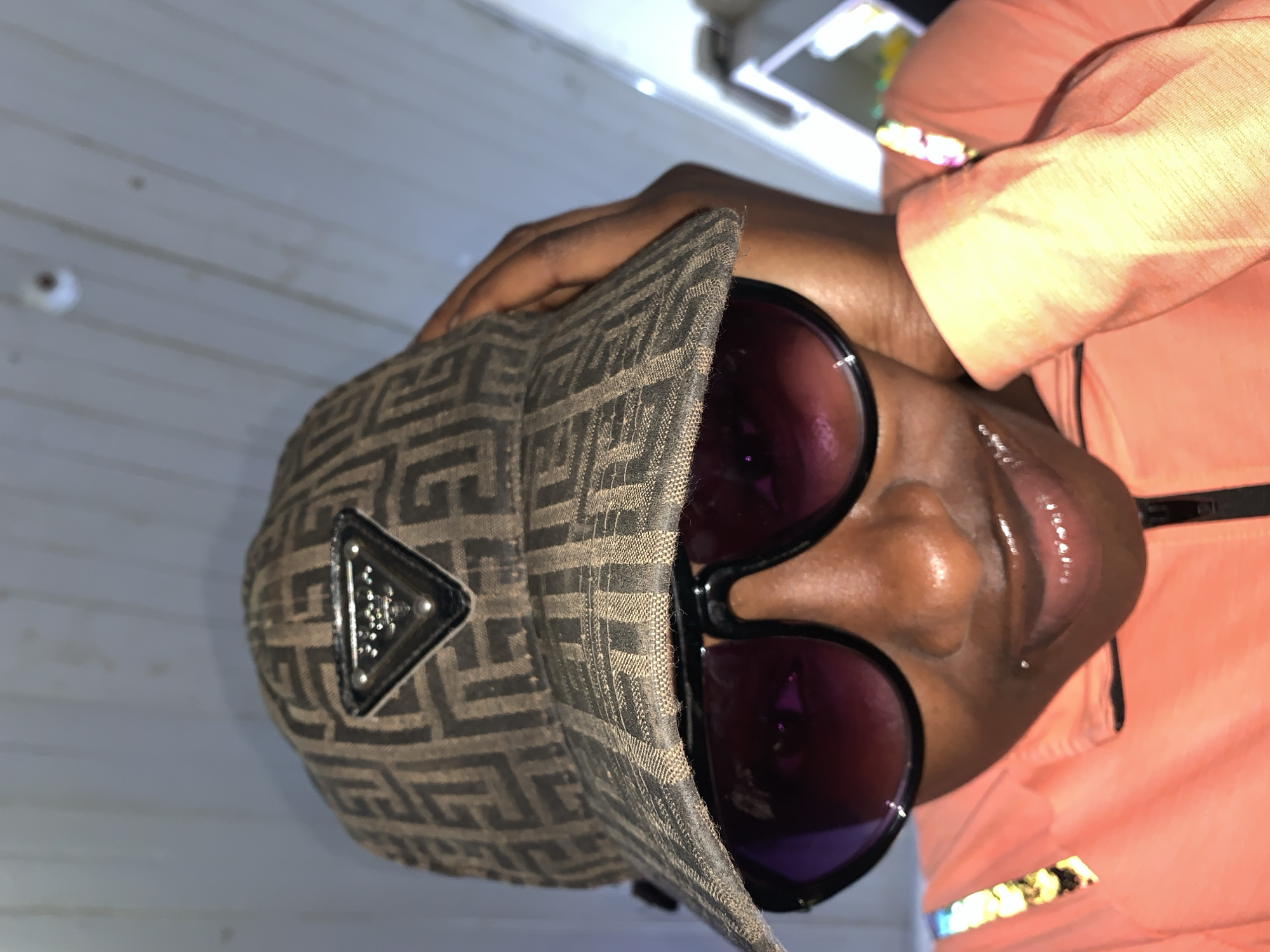 A person wearing a geometrically patterned baseball cap with a triangular emblem on the front and large, round sunglasses with purple lenses is visible. The person is also wearing an orange shirt or jacket. The background features a gray wall with horizontal planks, suggesting an indoor setting. The lighting is somewhat dim. The location is specified as New Stadium, Nigeria, without further detail about the immediate environment within the city. The image is taken from a close-up perspective, focusing on the face and upper body of the individual.
