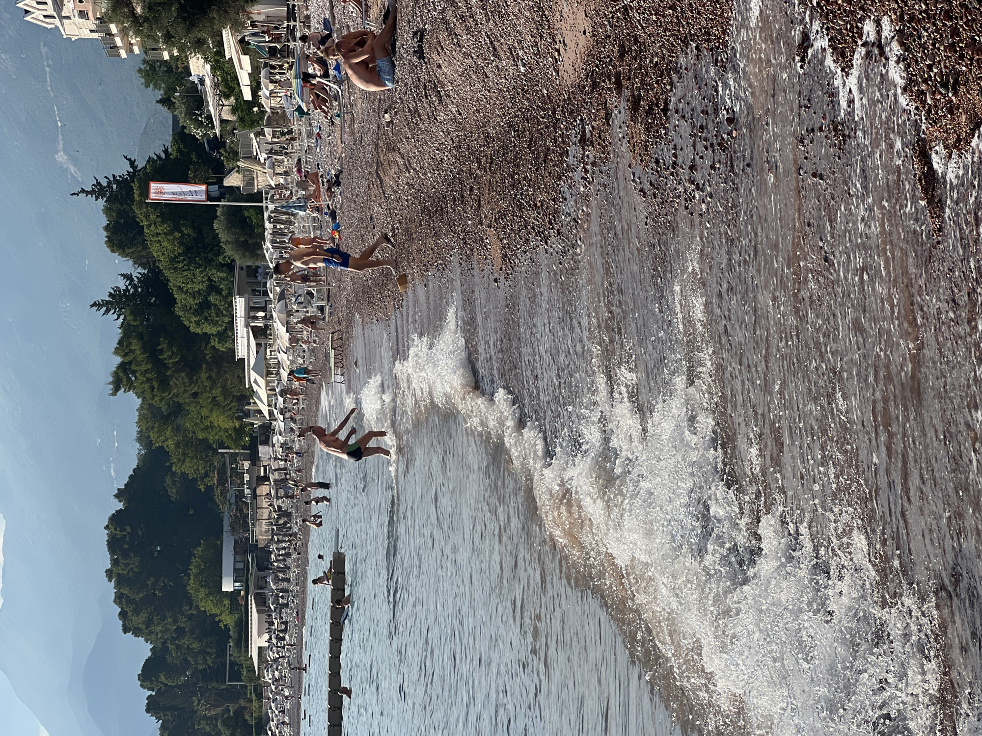 The image depicts a sunlit beach scene in Sveti Stefan, Montenegro. The foreground is dominated by the shallow, clear water of the Adriatic Sea gently lapping onto a pebble-strewn shore. Small waves create white foam as they break. In the middle ground, several individuals are visible. A man wearing dark swim trunks is partially submerged in the water, appearing to walk towards the shore with his arms extended. Further in the water, a young person in blue swim trunks stands. On the beach, other beachgoers are seen relaxing on lounge chairs or walking. The background reveals a bustling beach resort area with numerous white umbrellas providing shade, buildings nestled amongst lush green trees, and distant, hazy mountains under a clear sky. The overall impression is one of a vibrant summer day at a popular coastal destination.