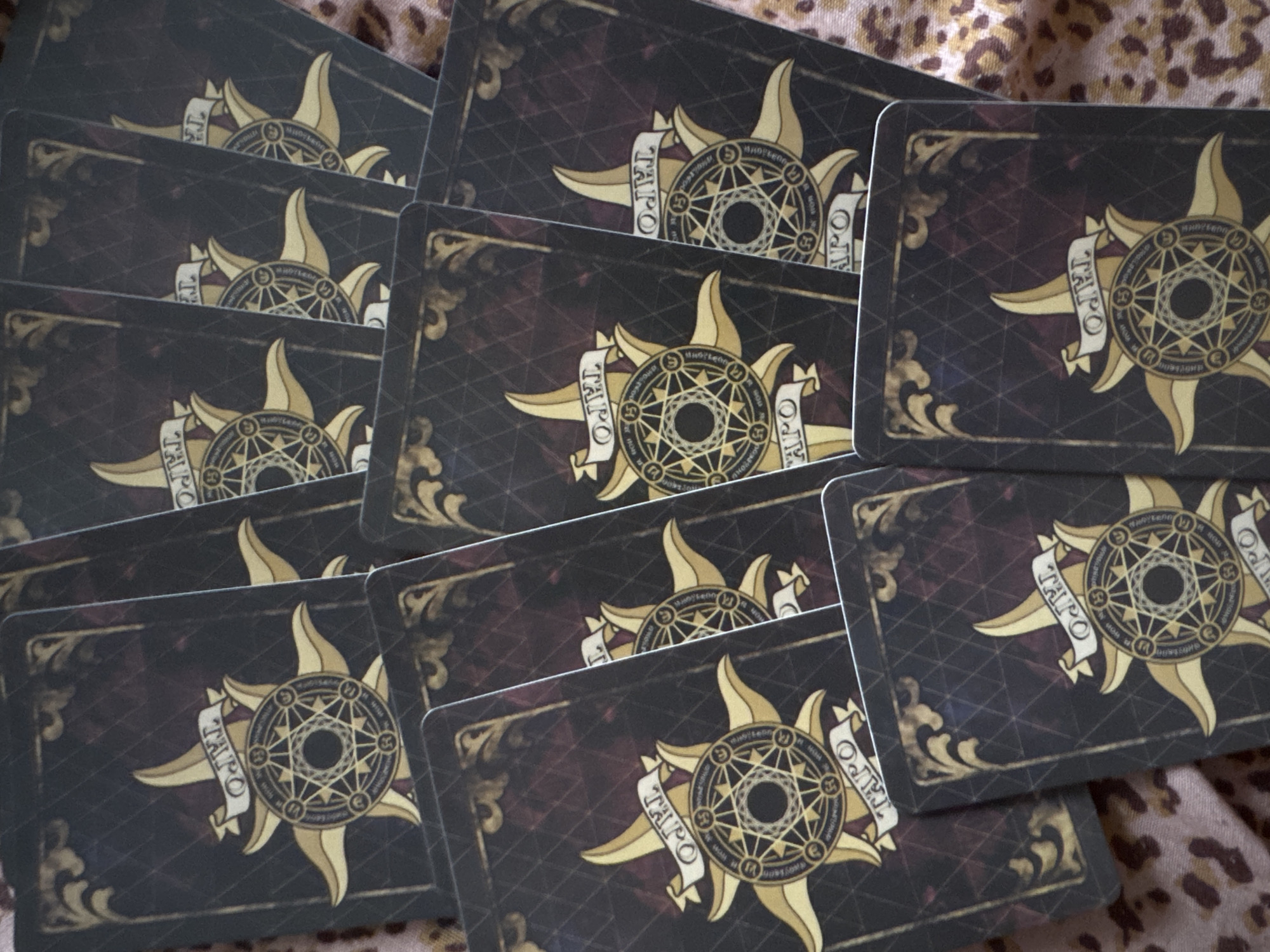 This image displays a collection of tarot cards, likely a deck, spread out on a surface. The cards feature a dark, ornate design with a central sun-like symbol radiating outwards. This symbol is surrounded by a circular pattern containing what appears to be astrological or alchemical symbols, and a banner with the word "TAROT" inscribed in gold lettering. The background of the cards is a deep, textured color with a subtle grid pattern and decorative scrollwork in the corners. The cards are overlapping and arranged in a casual, fanned-out manner, suggesting they are being used for a reading or are simply stored. The lighting is natural and evenly distributed, with no strong shadows indicating a specific time of day or weather conditions. The surface beneath the cards has a leopard print pattern, adding a contrasting texture and color to the scene. There are no people visible in the image. The overall impression is one of mystique and arcane symbolism. The image does not provide any specific location cues that would pinpoint it to Izhevsk, Russia, beyond the provided context.