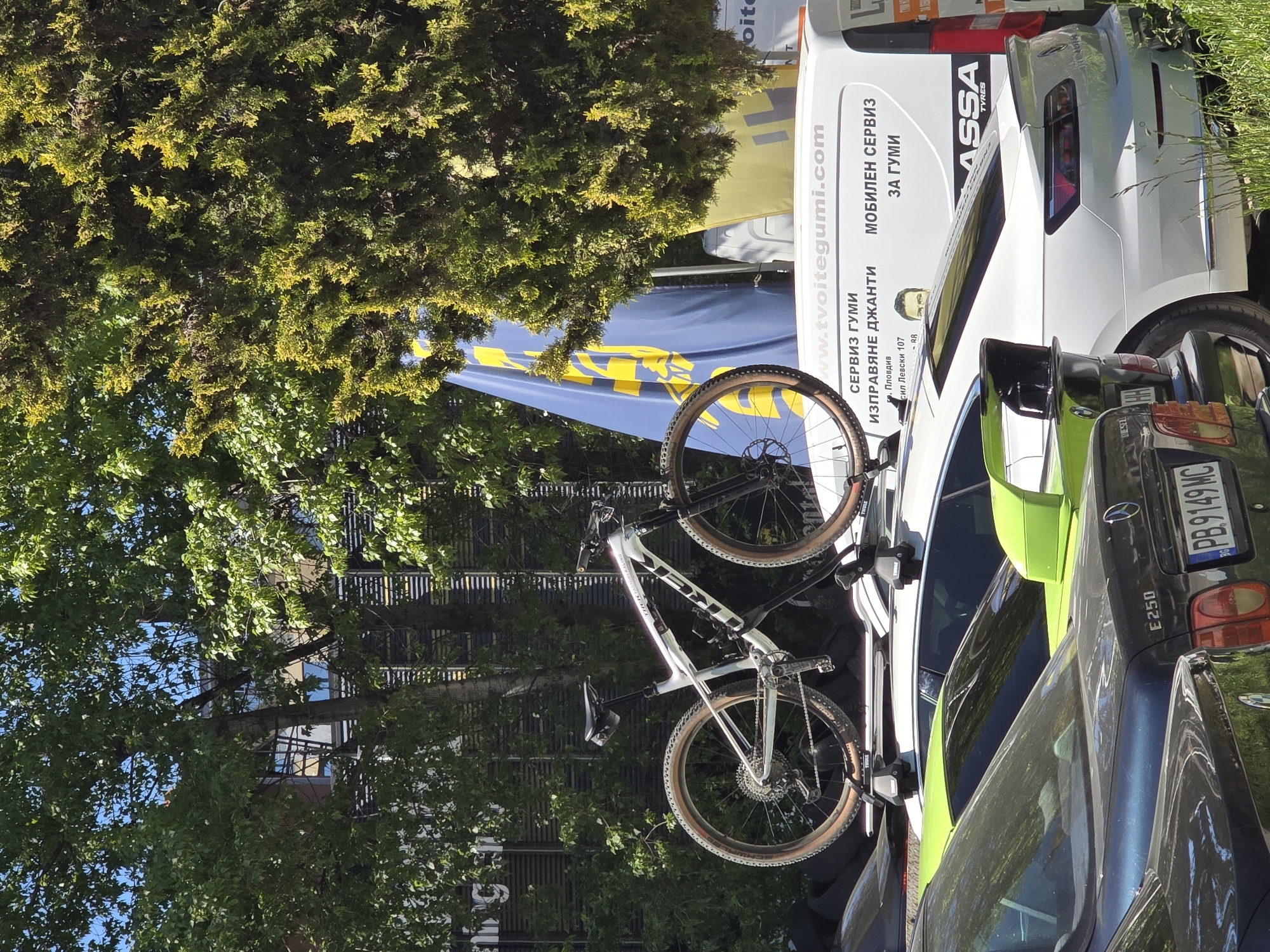 Here is a detailed description of the image:
**Scene Overview:**
The image shows a parking area outdoors, with several vehicles and trees visible.
**Vehicles:**
* A dark blue/black Mercedes E250 is parked in the foreground. Its license plate reads "PB.9149 MC".
* Next to the Mercedes is a vehicle with a lime green stripe running across the top.
* A white van with the text "www.tvoitegumi.com" visible on its side. Additional text includes "СЕРВИЗ ГУМИ" (SERVIZ GUMI), "ИЗПРАВЯНЕ ДЖАНТИ" (IZPRAVYANE DZHANDI) and "МОБИЛЕН СЕРВИЗ ЗА ГУМИ" (MOBILEN SERVIZ ZA GUMI). The word "ASSA TYRES" can also be seen.
* The back of a white car is visible behind the van, with the word "oite" visible on the van.
**Other Objects:**
* A mountain bike is mounted on a roof rack on the white vehicle.
* A large blue and yellow advertising banner is behind the van.
**Environment:**
The setting is urban, as evidenced by the presence of parked cars and a building partially visible through the trees.
**Location:**
The Cyrillic text on the van suggests a location where Bulgarian is spoken. The mention of the city "Пловдив" (Plovdiv) and "бул. Левски 107" (Levski Blvd. 107) suggests that the image was taken in Plovdiv, Bulgaria, at or near 107 Levski Blvd.