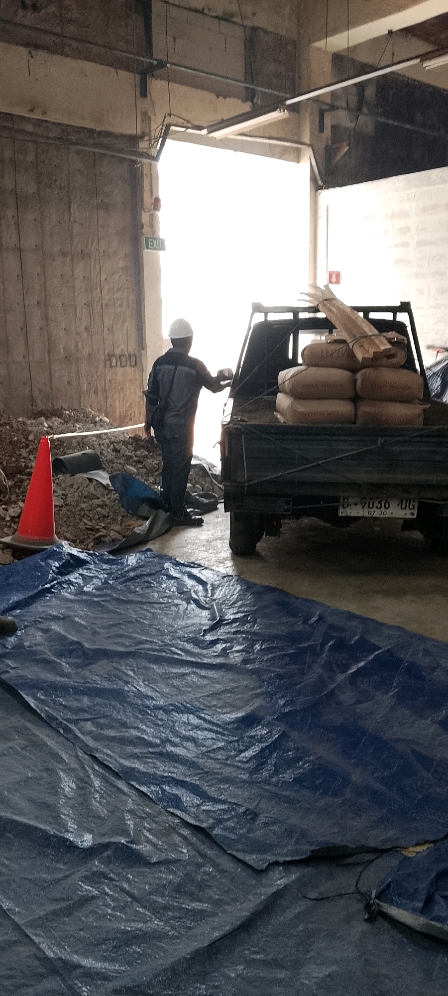 A construction worker wearing a hard hat and dark work clothes stands near the back of a pickup truck laden with sacks of cement and wooden planks. The truck is parked in what appears to be a covered area, possibly a warehouse or under an overpass, with a brightly lit opening visible in the background. Debris, including rocks and a red traffic cone, is scattered on the ground around the worker and the truck. A large blue tarp covers a portion of the foreground. The license plate on the truck, visible in the lower right, reads "B-9036 UG". The scene is located in Depok, Indonesia.