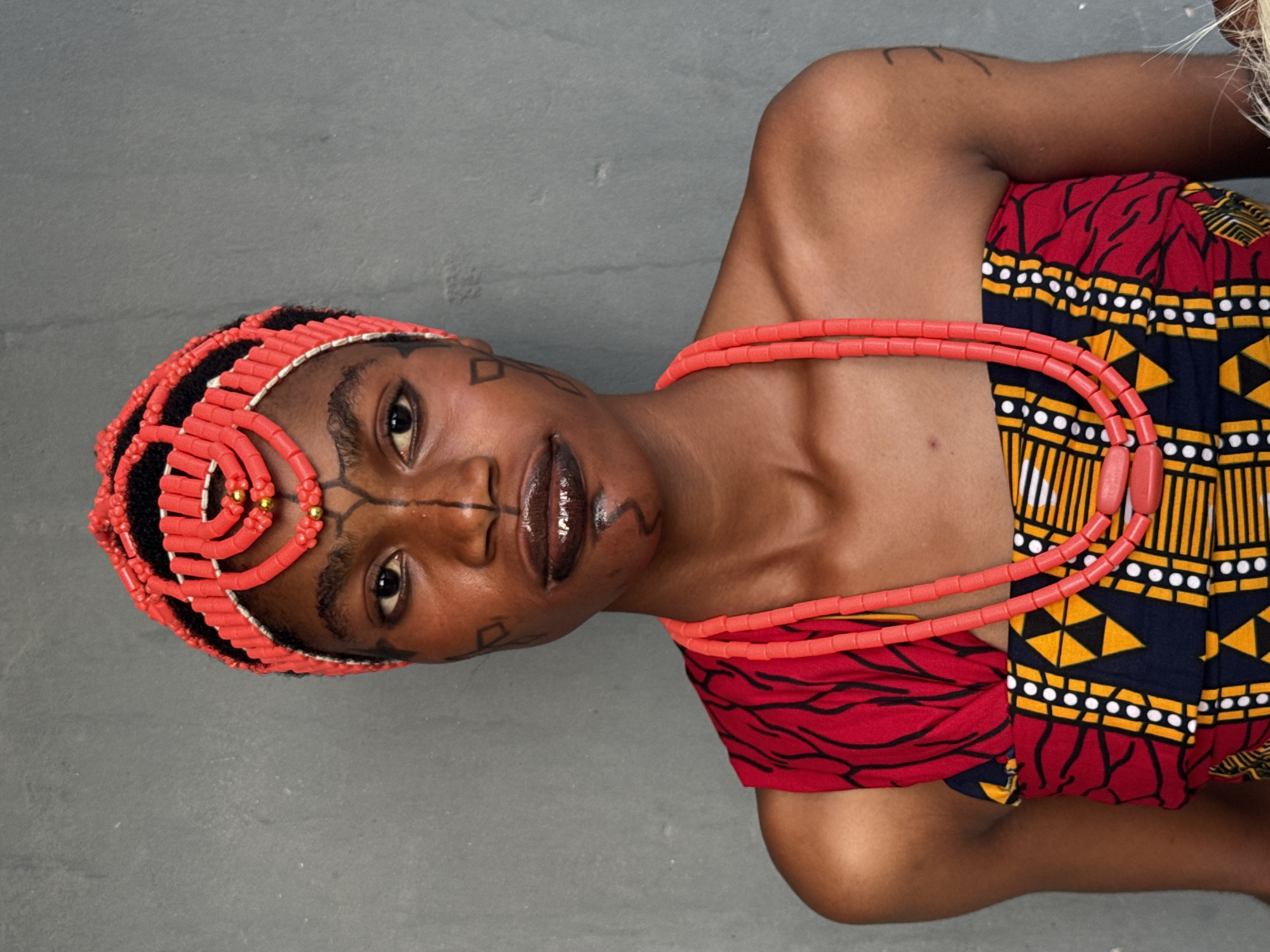 A woman is facing the camera with her head turned slightly to the left, wearing a red beaded headdress, necklace and a patterned red, gold, white, and blue dress. She has facial markings on her forehead and cheeks. She is in a studio. There is no evidence to prove the location is the city of Badagry, Nigeria.
